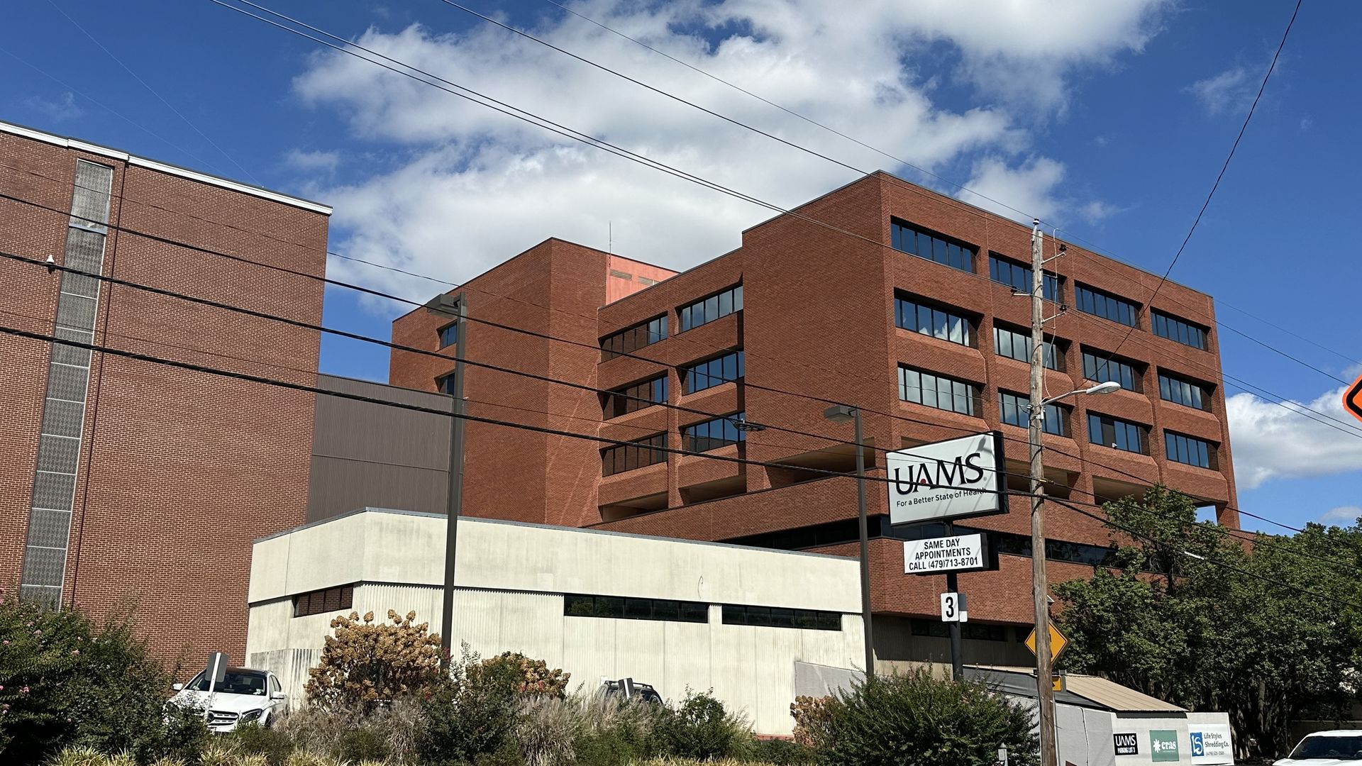 Blue sky with white clouds above a red brick office building; overhead power lines cross the scene, with a UAMS sign and a few parked cars near the street corner.