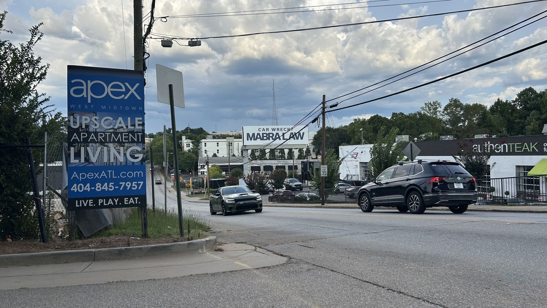 Street scene with a large blue sign for Apex West Midtown upscale apartments, cars on the road, and a billboard advertising Mabra Law car wreck law firm under a cloudy sky.
