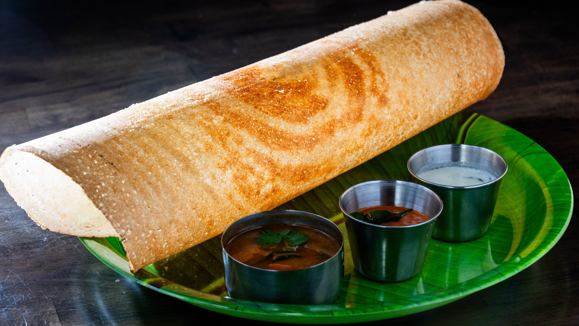A large rolled dosa on a green plate with three small cups of dipping sauces.