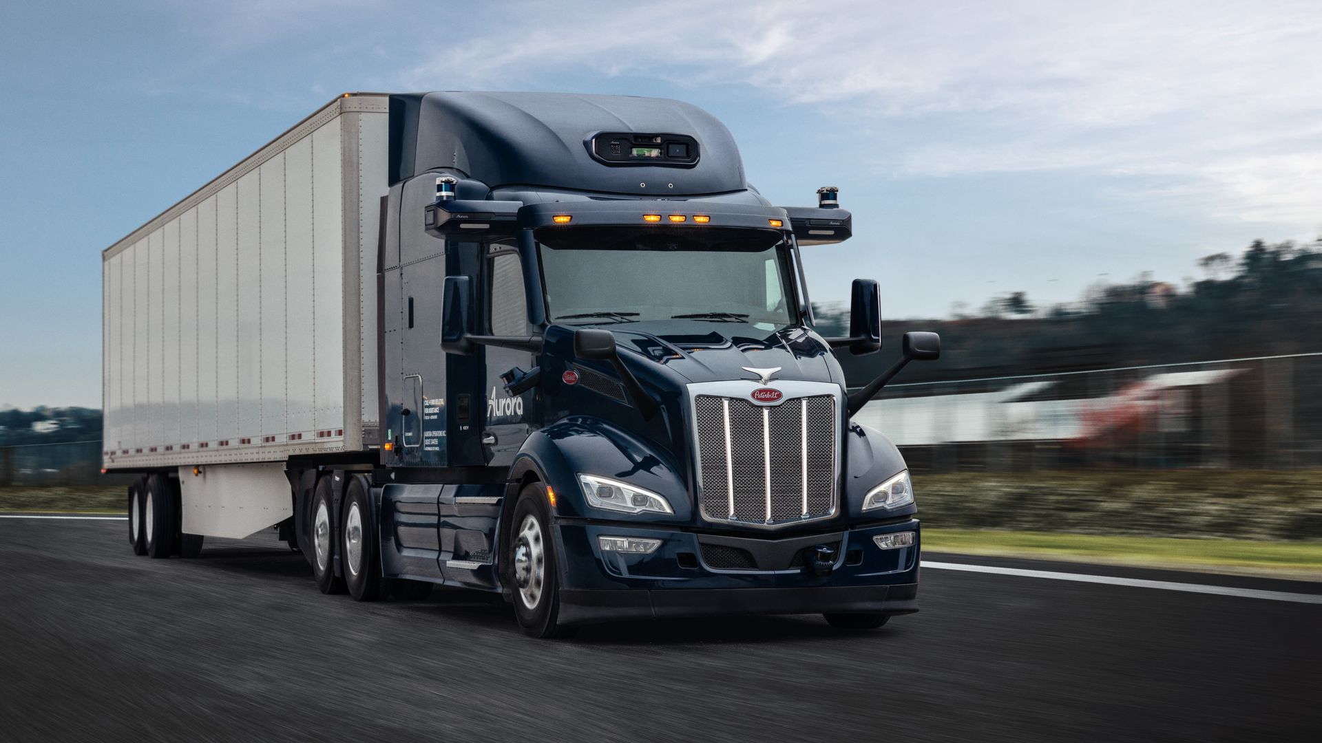 Dark blue semi-truck with a white trailer driving on a highway against a cloudy sky, featuring the logo "Aurora" on the door and glowing orange lights above the windshield.