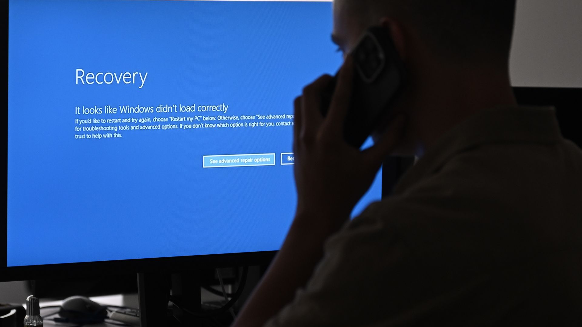 A person holds a phone to their ear while sitting in front of a computer. The computer has a blue screen with a "recovery" message on it. 