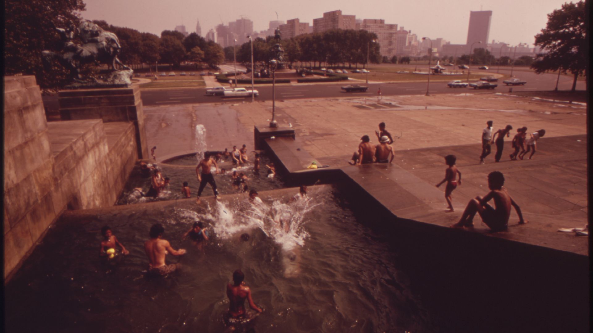 Public fountain and shallow pool with kids splashing; people sit on the edge; a wide plaza and road with cars, and a distant city skyline.