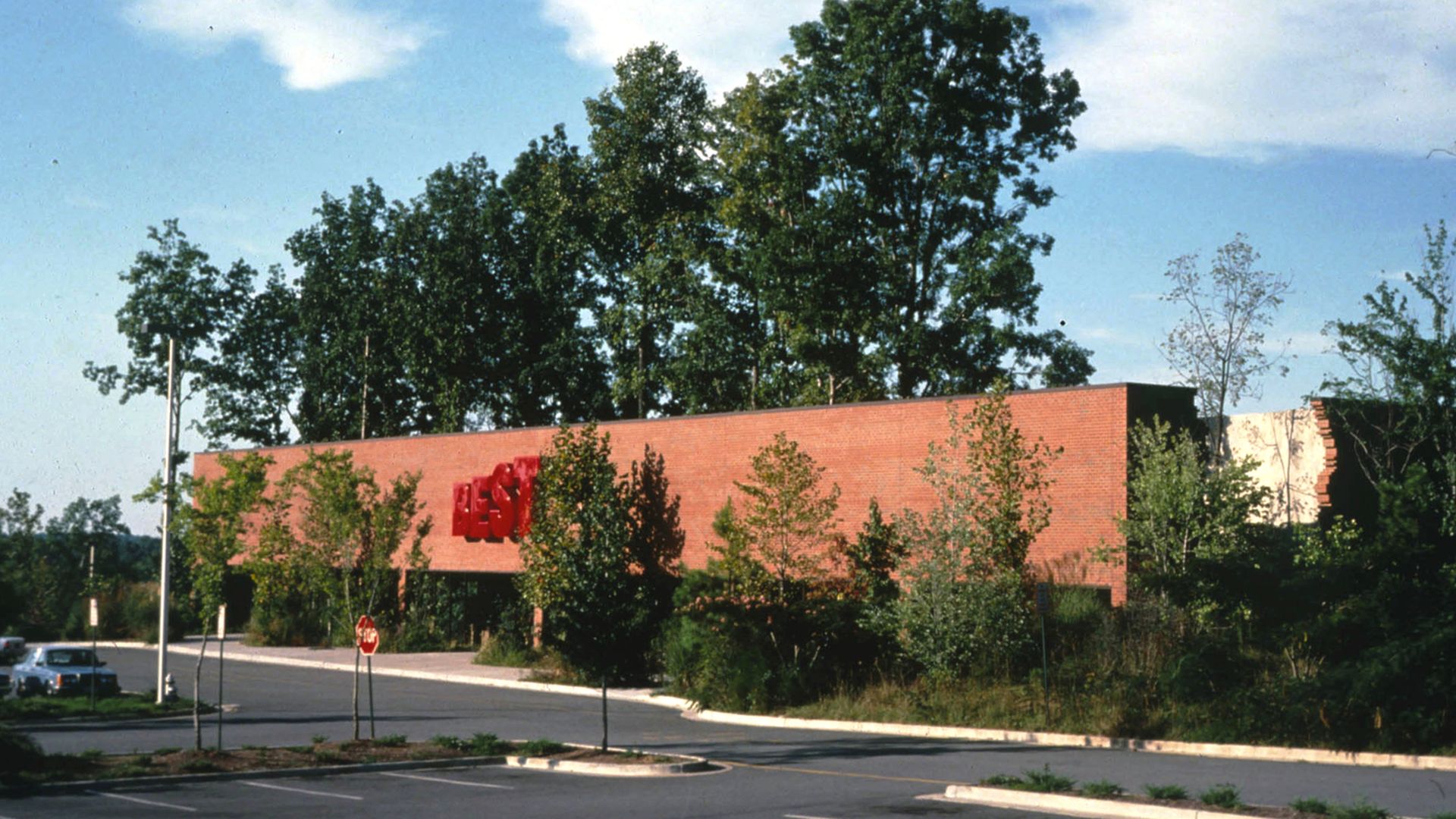 A long, low brick building with a large red sign on its front wall sits beside a parking lot, surrounded by trees and shrubs under a bright blue sky.