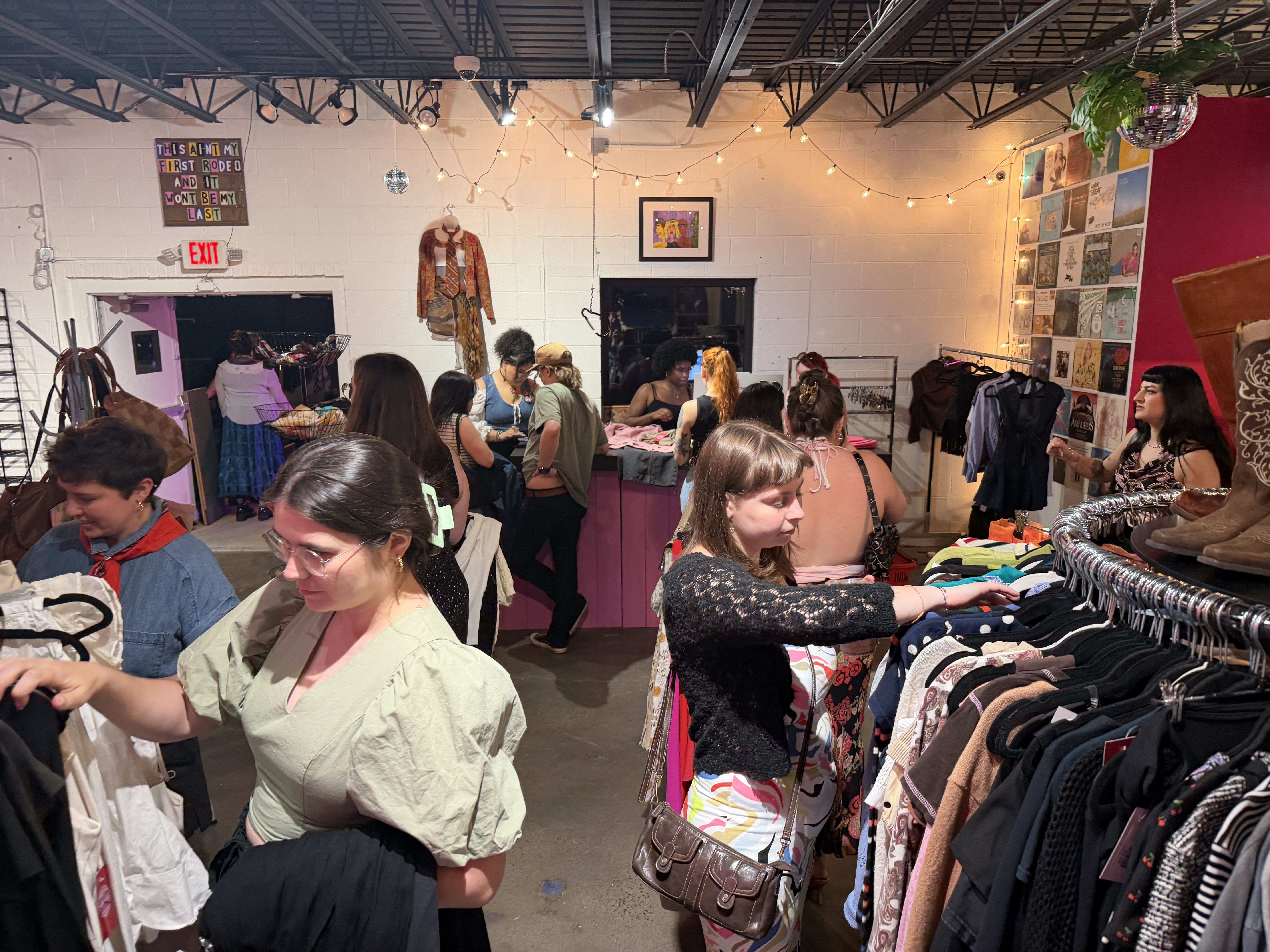 Busy thrift store with shoppers browsing racks of colorful clothing. Warm string lights, a pink counter, a hanging mannequin, wall art, and a disco ball adding a cozy, retro vibe.