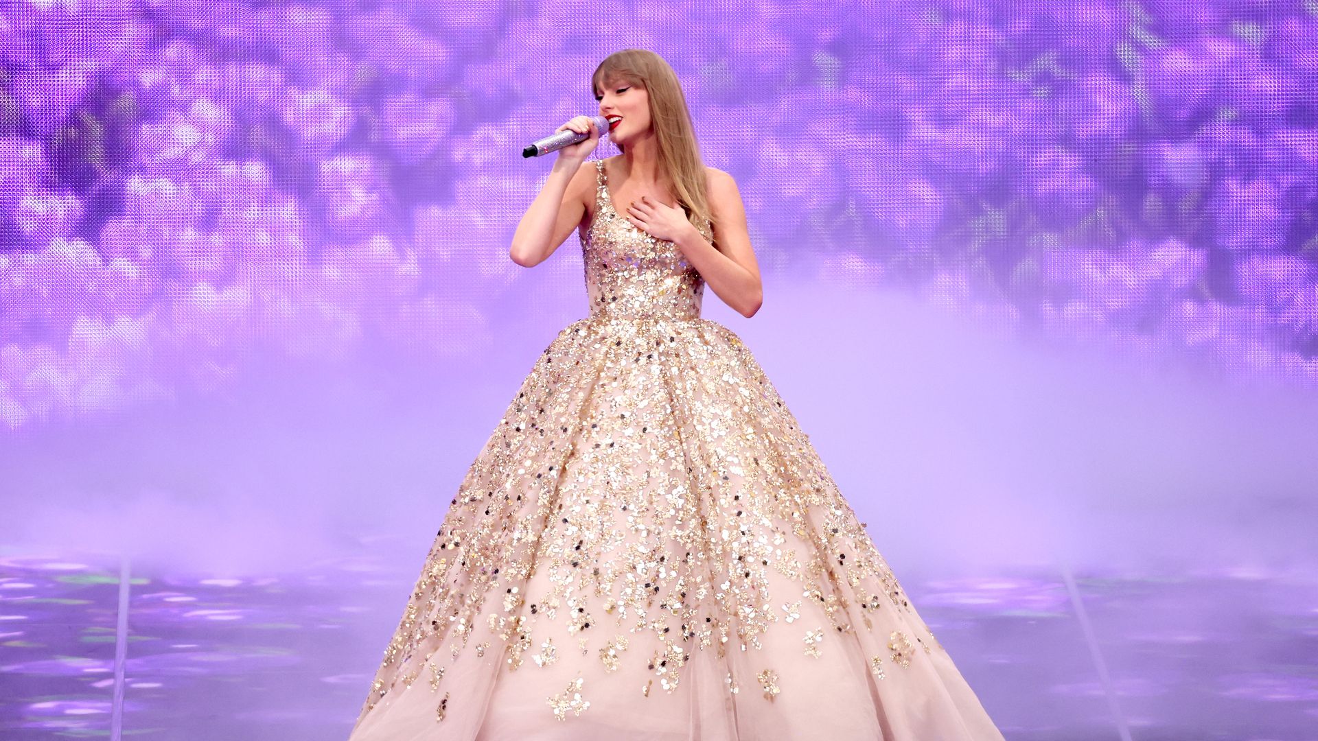 Female singer in a sparkling pale pink full-skirt gown with gold sequins performs on a purple-lit stage, holding a silver microphone and touching her chest as fog swirls behind her.
