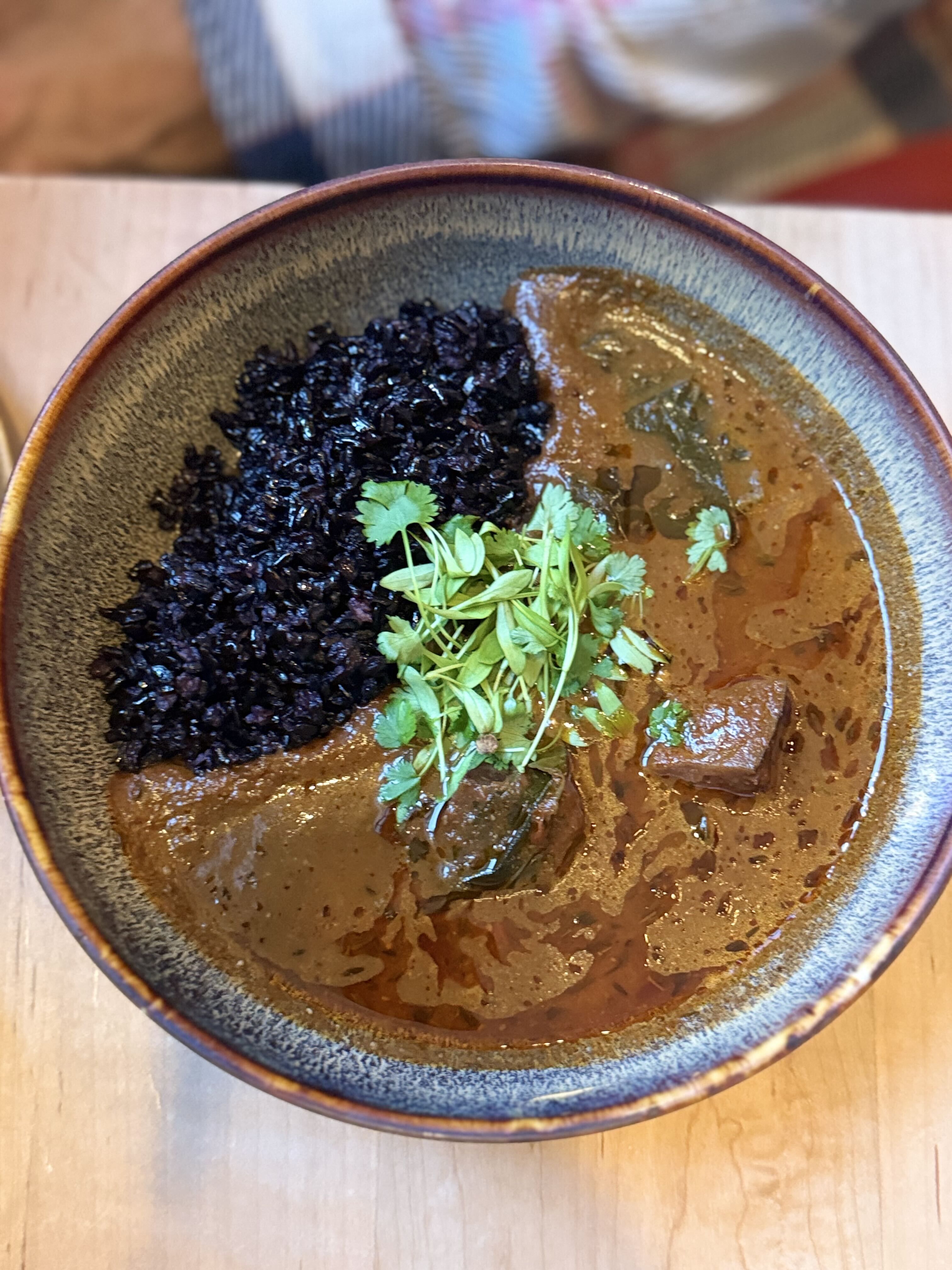 Top-down view of a ceramic bowl with black rice on the left and a brown curry with meat on the right, topped with fresh cilantro sprouts; a wooden table is visible underneath.