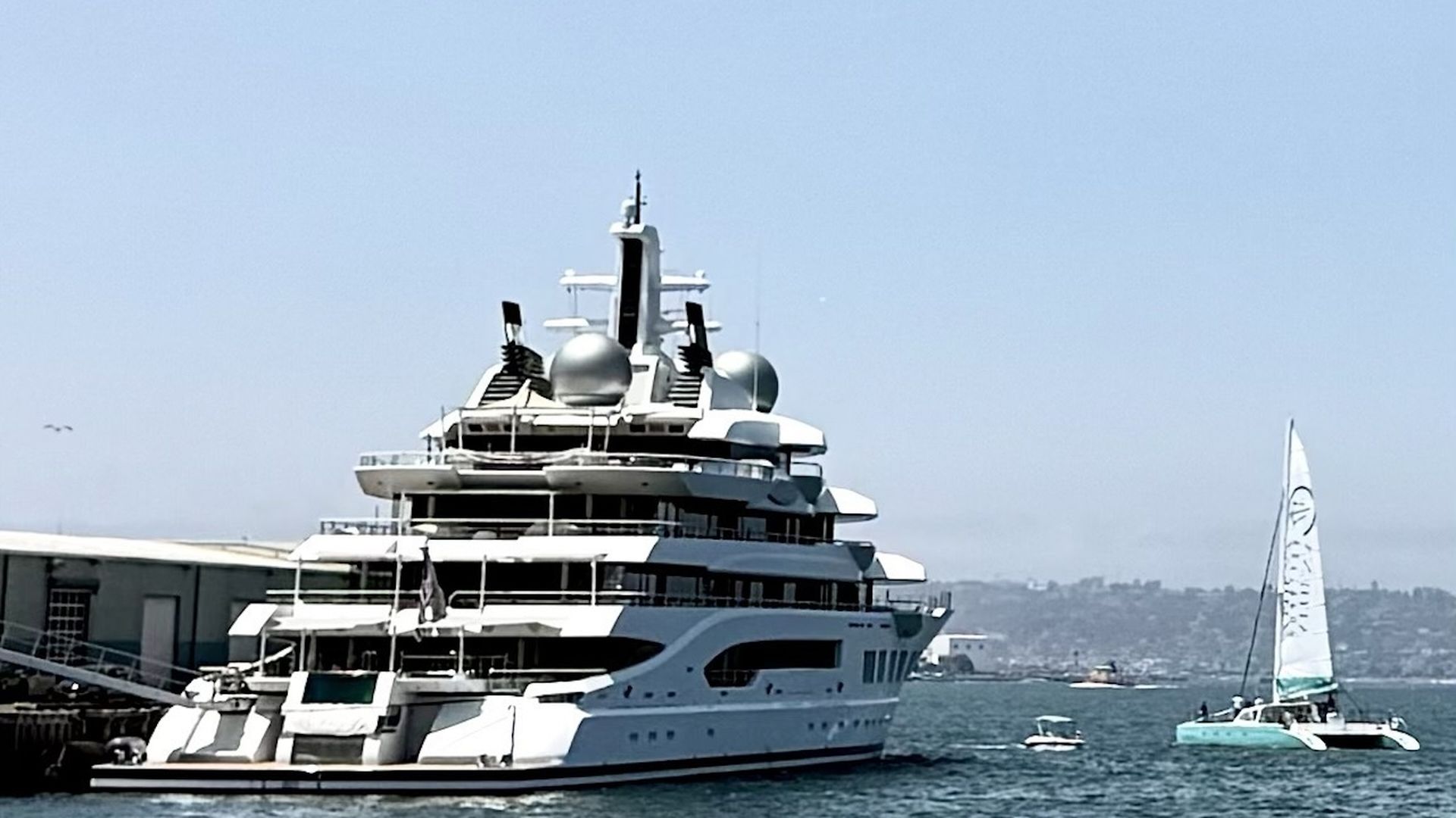 A large six-deck yacht docked in a harbor, being approached by a small, two-hull sailboat