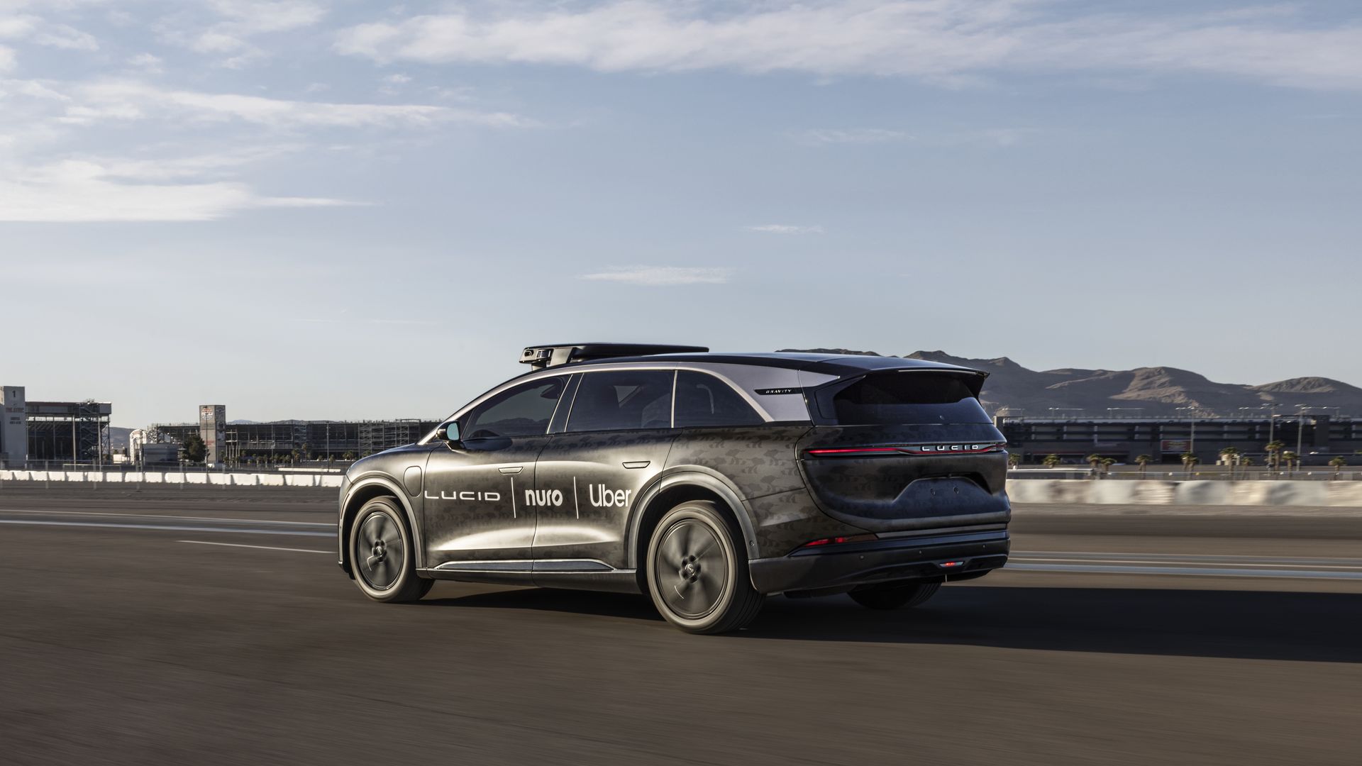 Black Lucid Gravity electric SUV with logos for Lucid, Nuro and Uber on the side, driving on a closed track with mountains in the background and blue sky above.