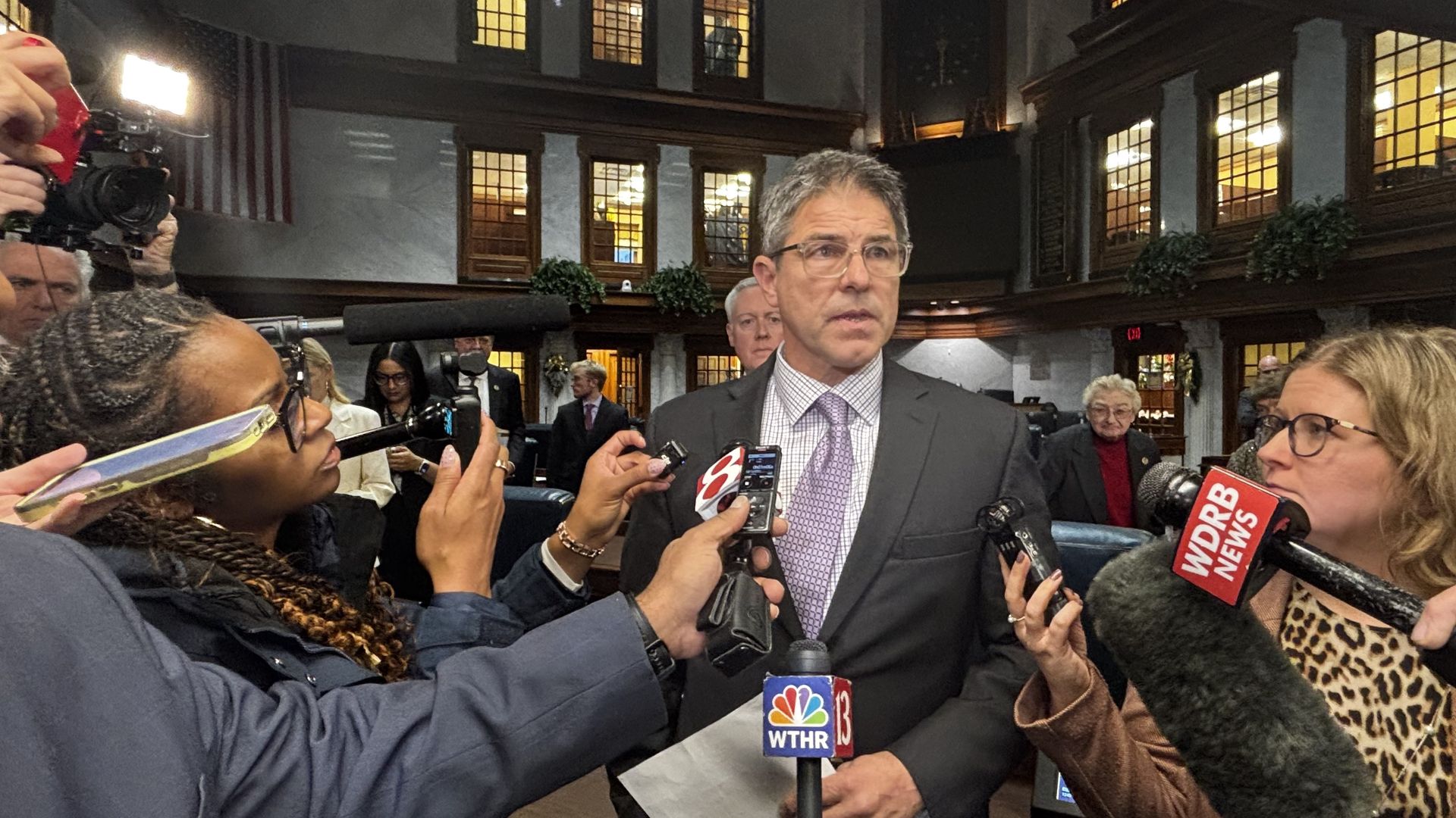 Man in glasses and suit speaking to reporters holding microphones labeled WTHR 13, WDRB News, and Channel 8 in a wood-paneled room with large windows and people in the background.
