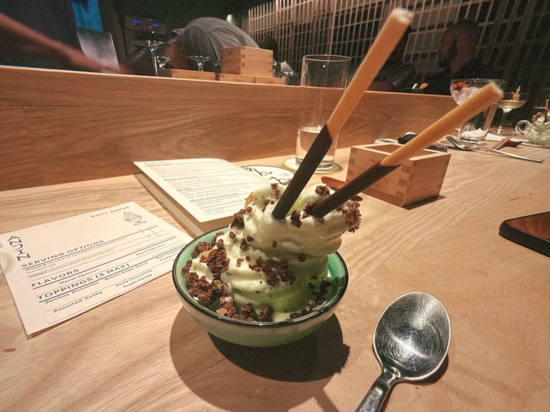 Cup of green matcha soft serve ice cream with chocolate sprinkles and two chocolate-dipped biscuit sticks, placed on a wooden table with a spoon and menus nearby.