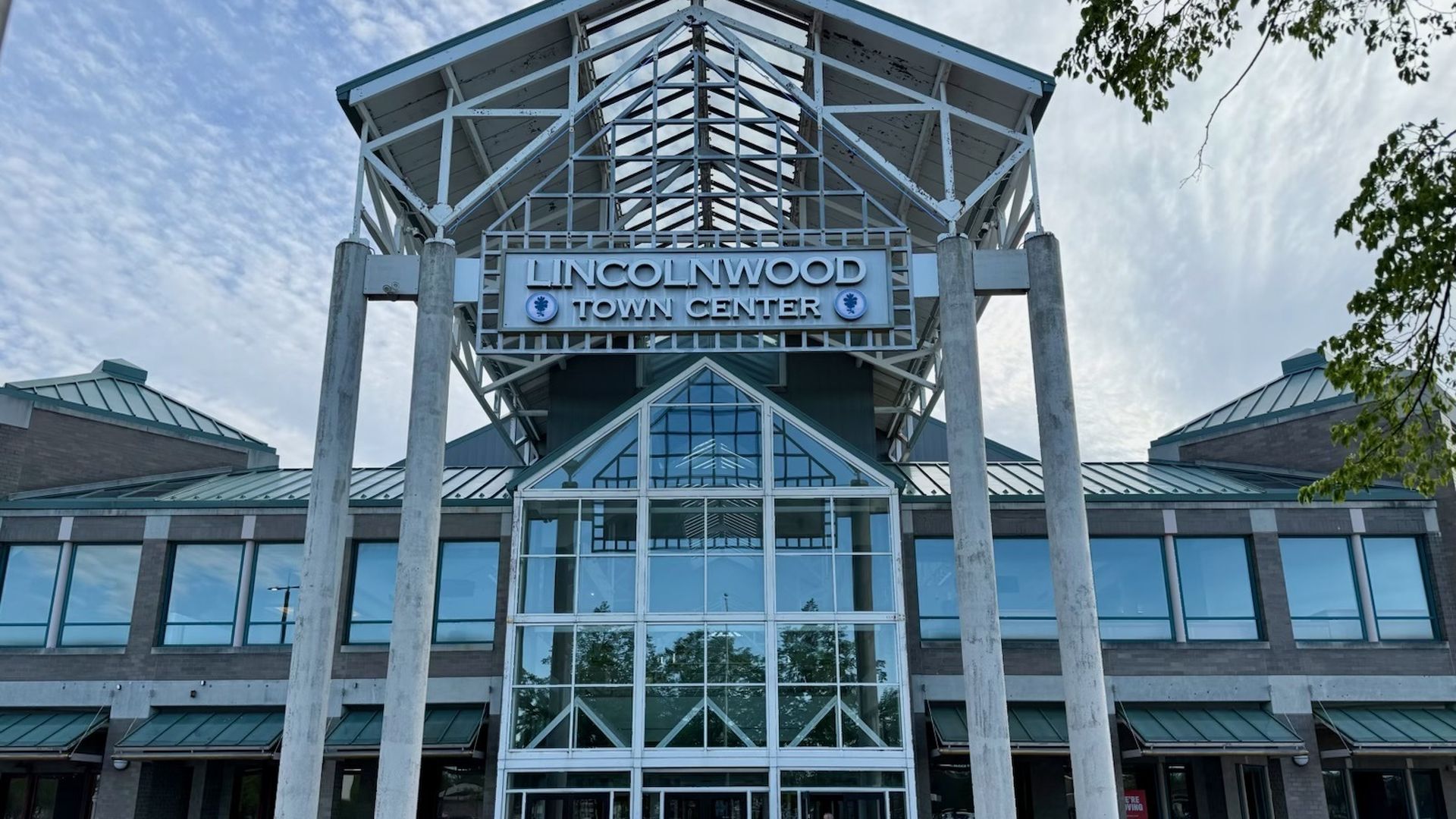 Front view of Lincolnwood Town Center: glass atrium, pale steel framing, and the sign reading "LINCOLNWOOD TOWN CENTER". Two tall concrete columns frame the entrance; a child in red stands on the left with blue sky above.