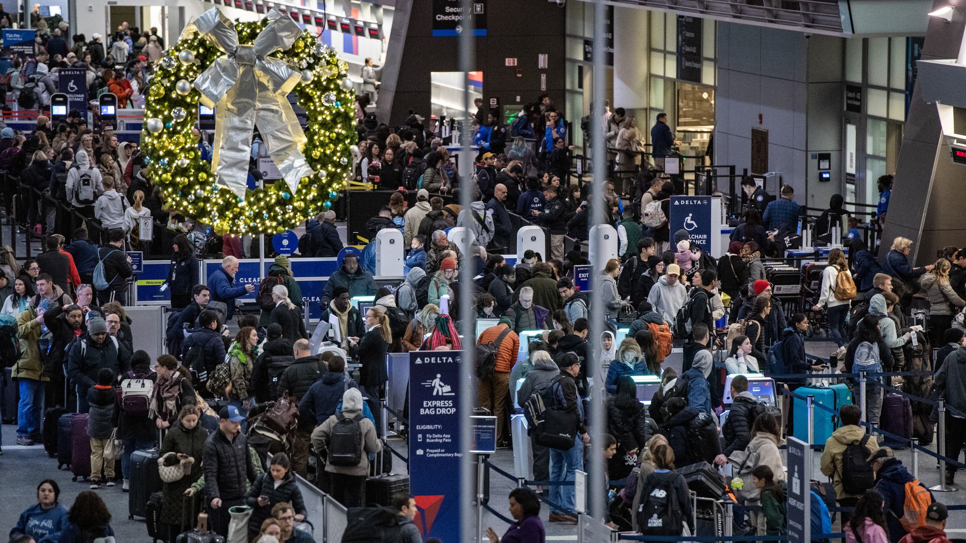 Crowded airport security checkpoint decorated with a large lit Christmas wreath and silver bow, with many travelers waiting in lines carrying luggage in winter clothing.