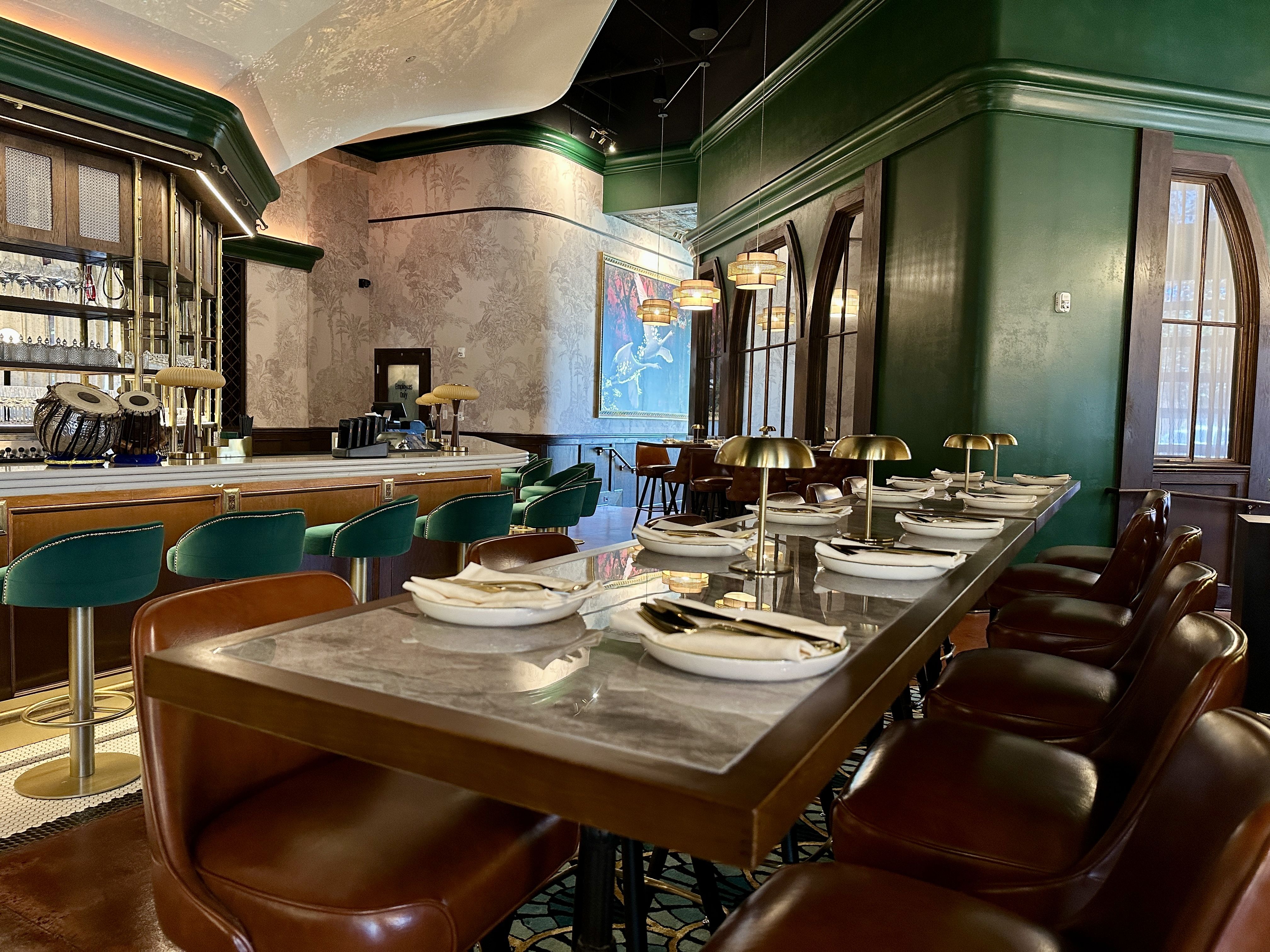 Elegant bar and dining area with dark green walls, gold-trimmed bar, marble countertops, brown leather stools, and round amber pendant lights. Tables set with white plates and napkins.