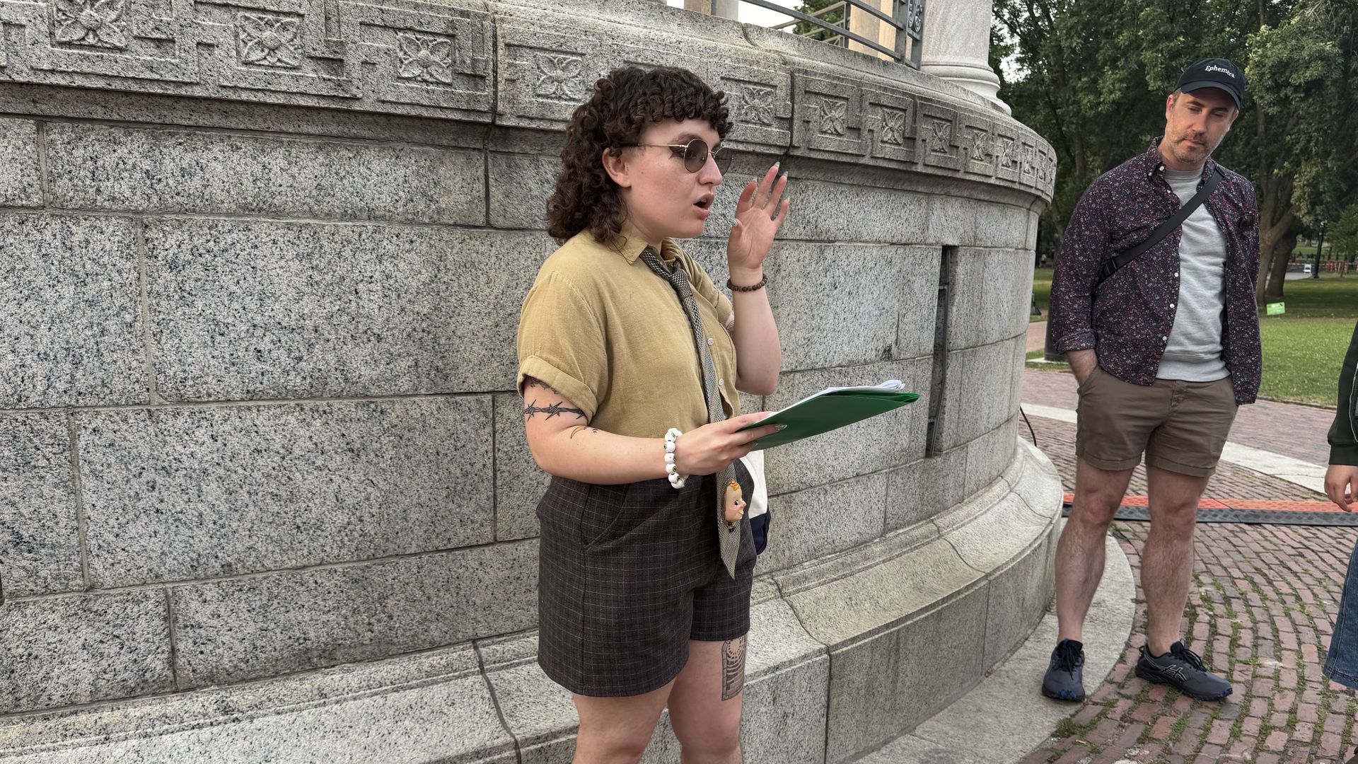 A tour guide with a tie that has a doll head stands in front of the Parkman Bandstand on the Boston Common, holding a folder with a script as he leads a group of tourists through a tour of LGBTQ+ history.
