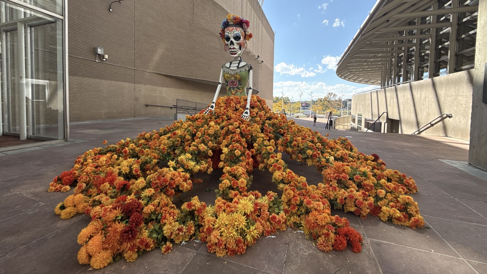 Large Day of the Dead figure with a skeleton figure wearing a flower crown and a dress made of orange and yellow flowers.