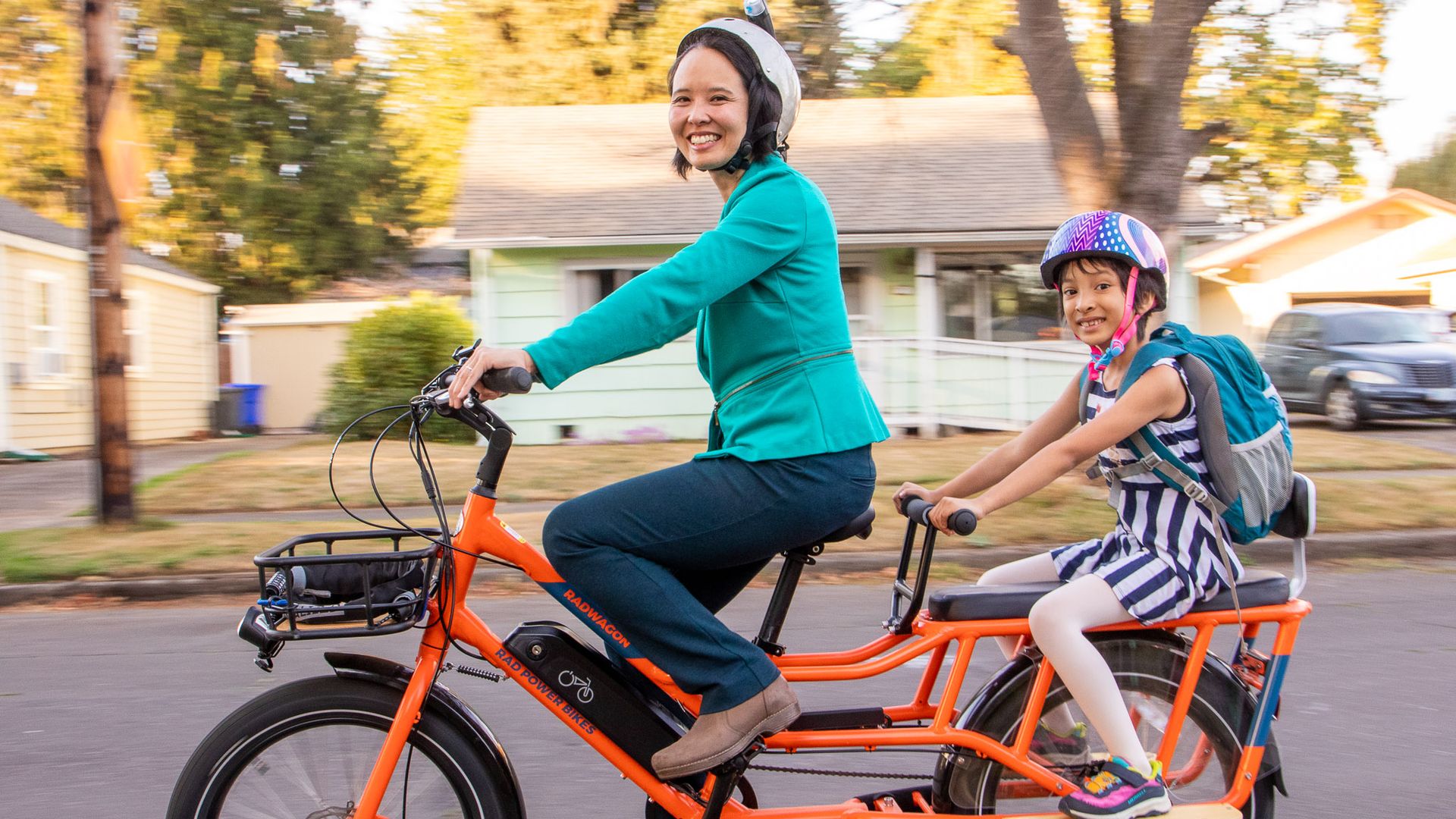 A woman riding a bright orange, long bike, with a child wearing a backpack on the extender seat in the rear.