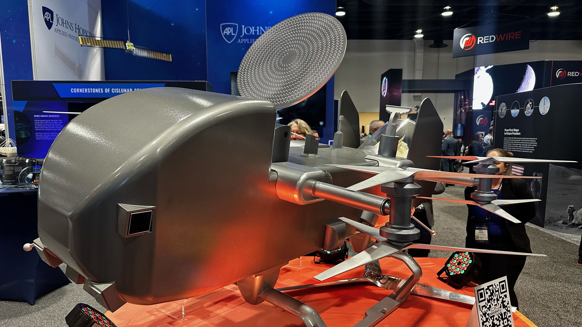 A large gray unmanned drone on a red exhibition platform, with multiple rotor arms and blades. Blue Johns Hopkins Applied Physics Laboratory booths and several attendees visible in the background.
