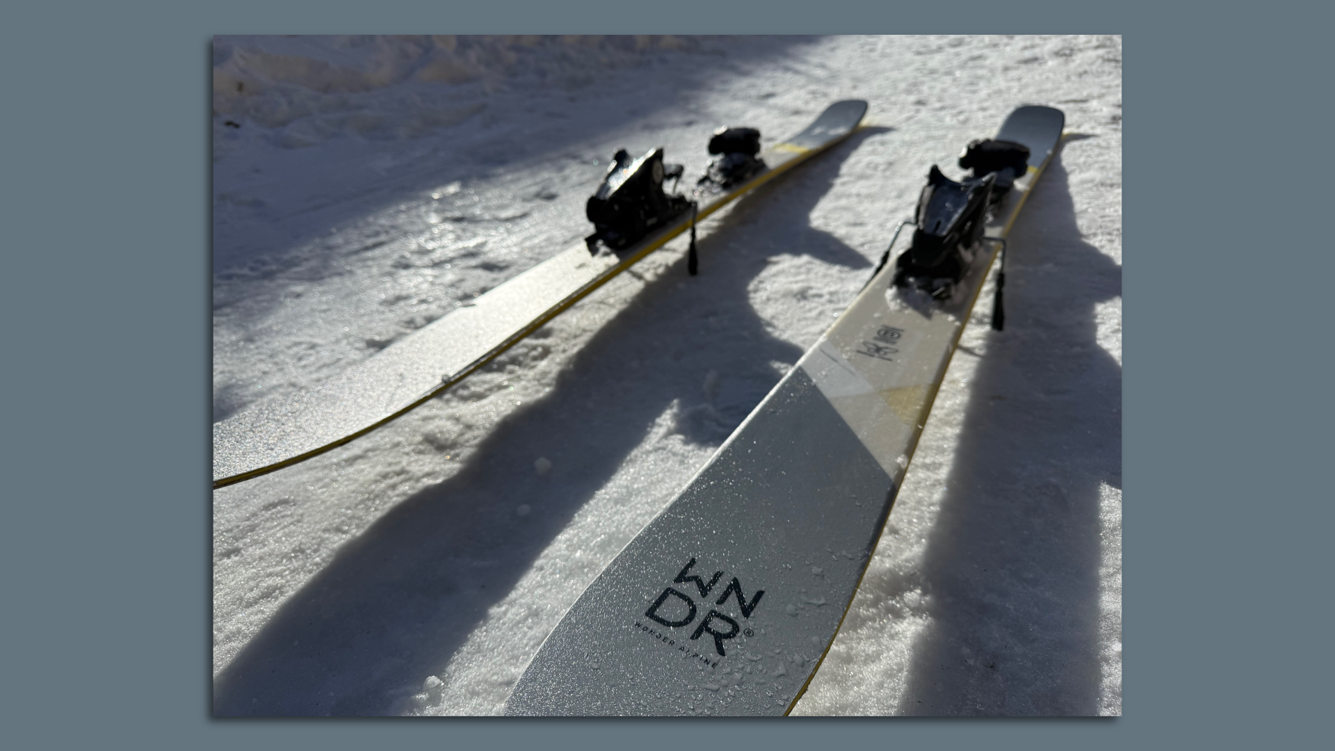 A close-up photo of gray and yellow WNDR Alpine skis, sitting on snow.