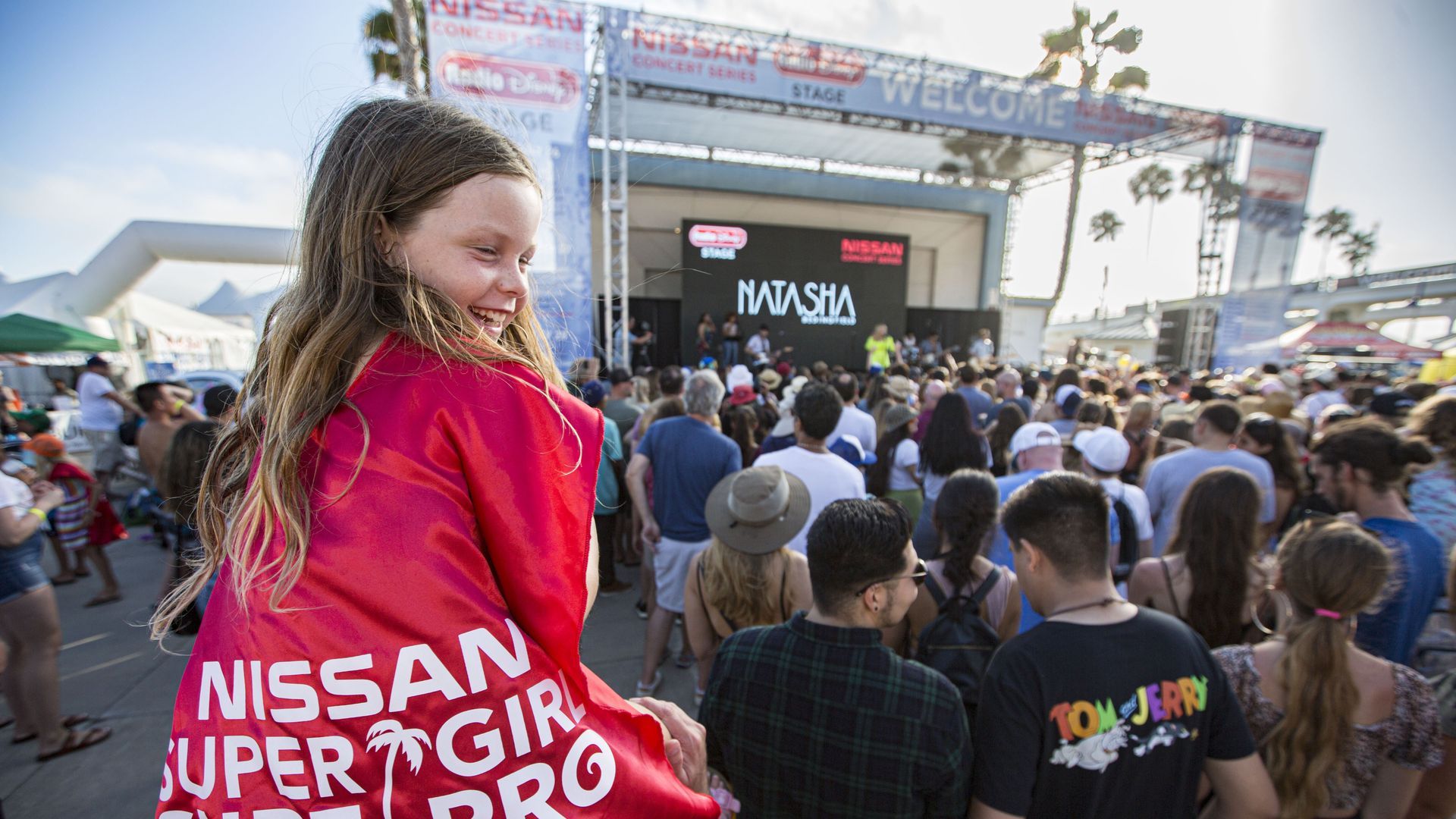 A girl wearing a red cape that says "Super Girl Surf Pro" sits on someone's shoulders at an outdoor concert by the beach.