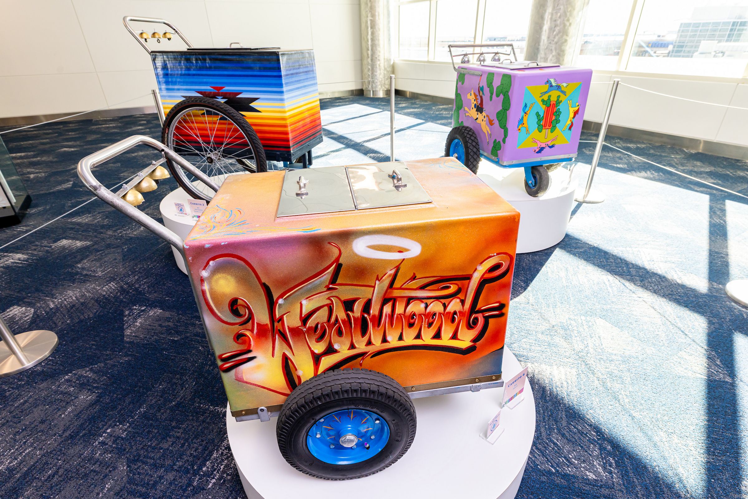 Three colorful hand-pulled carts displayed indoors on white platforms with artistic designs: orange one with "Westwood" graffiti, a blue striped one, and a purple one with animal and cactus artwork.