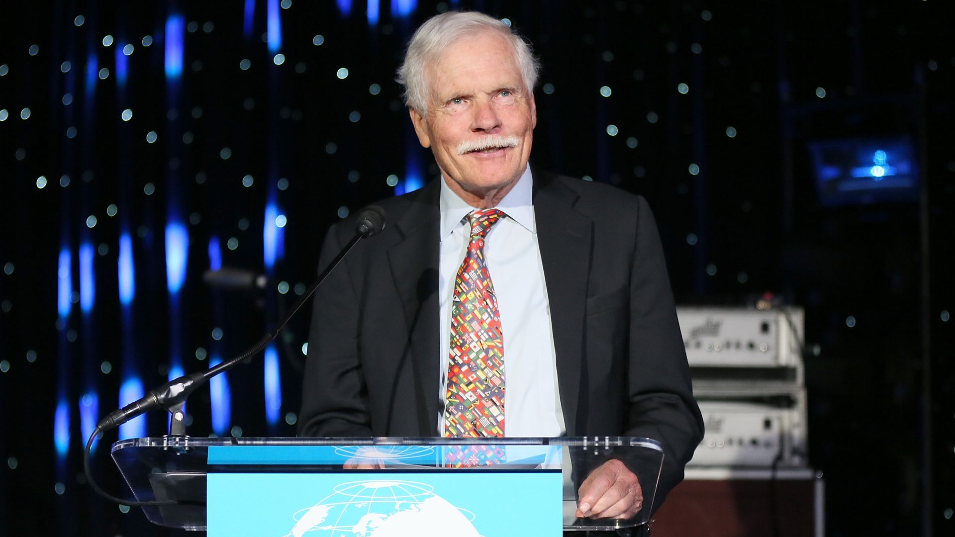 A man wearing a suit and tie covered in small colorful splotches speaks at a clear lectern with a blue sign on the front. TV screens tuned to the same image are in the background.