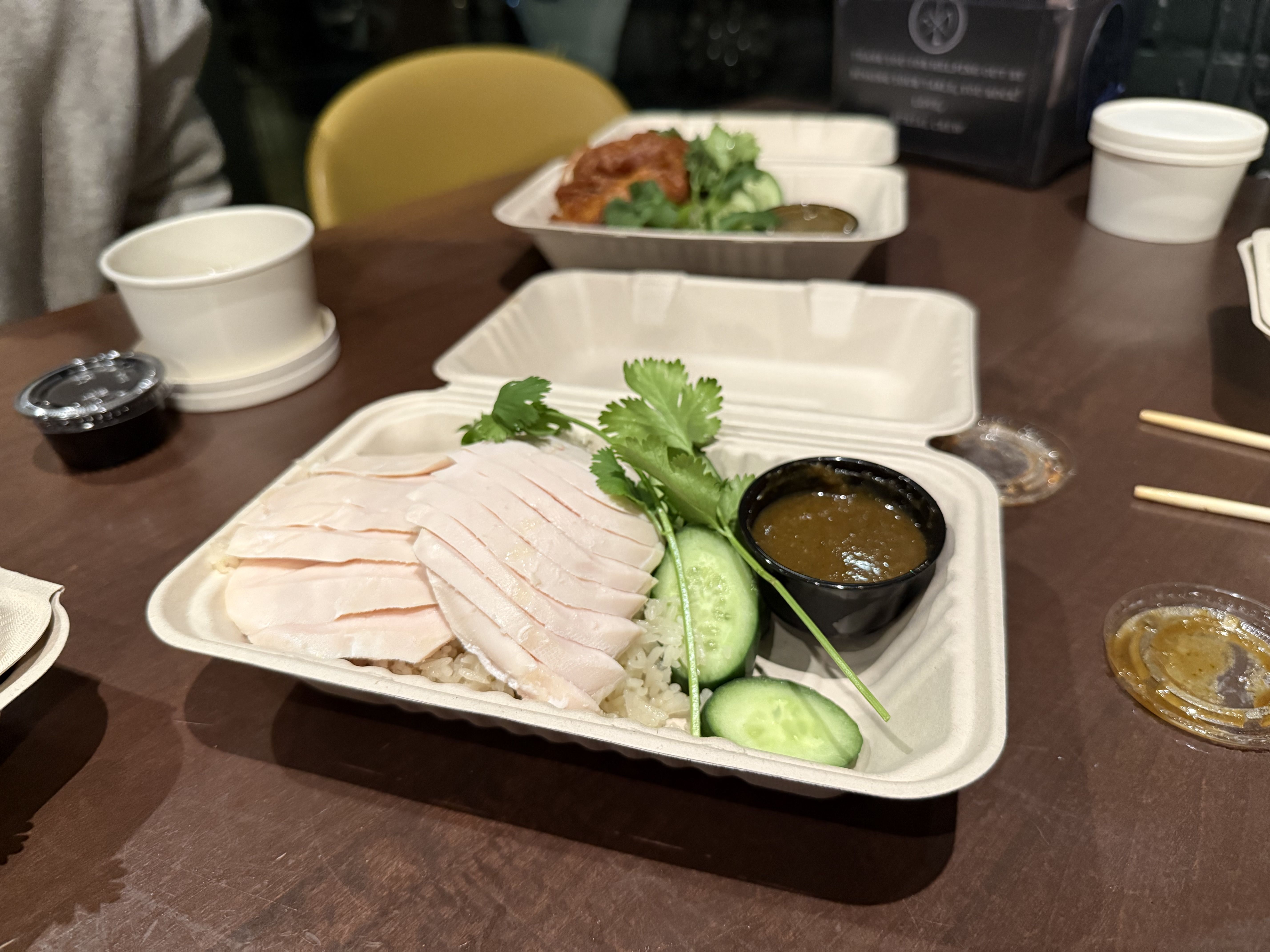 Takeout container with sliced chicken on rice, cucumber slices, cilantro, and a small cup of brown sauce, on a brown table with another container of food and chopsticks visible in the background.