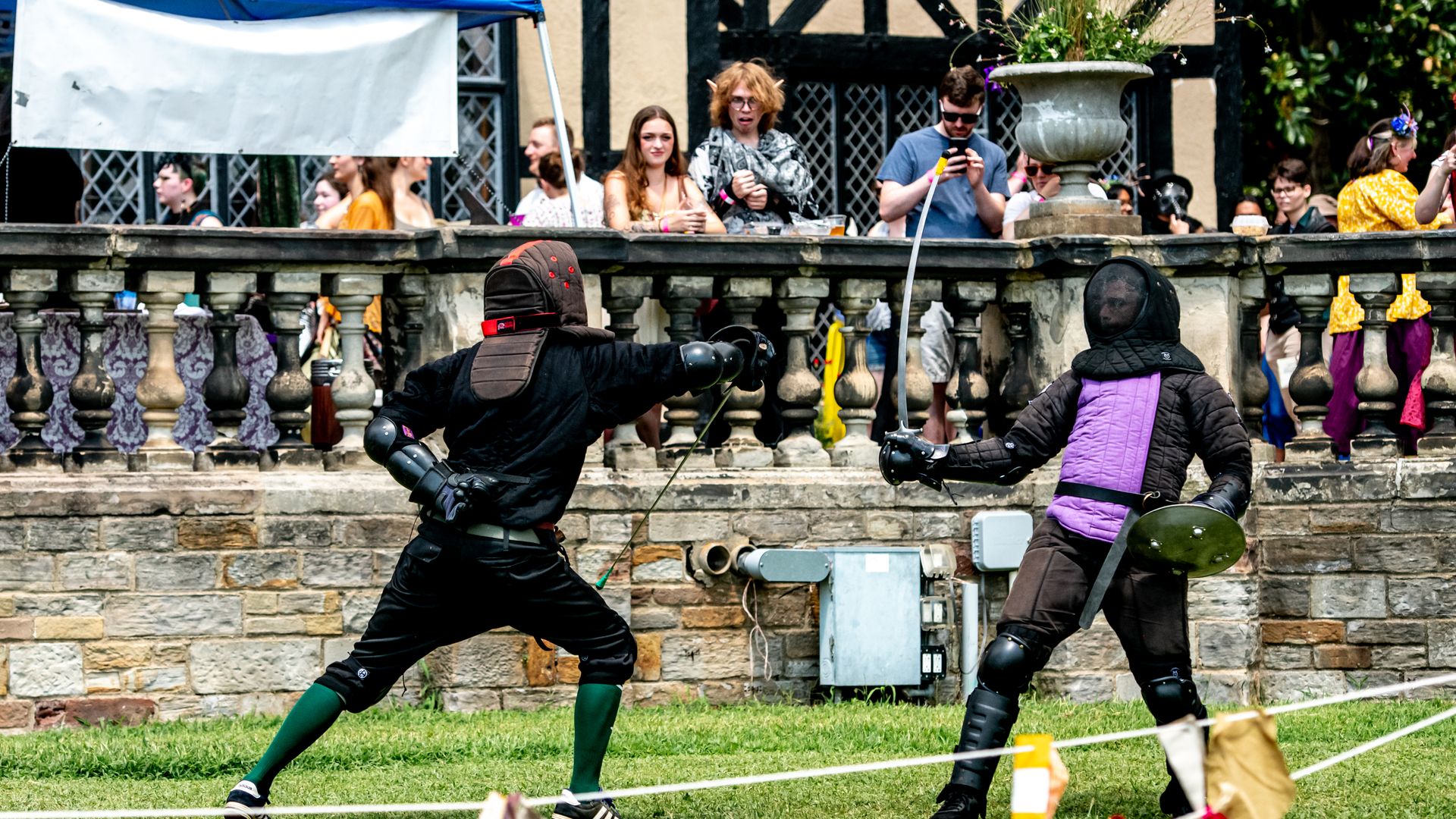 Two armored combatants duel with swords on a grassy area, one in black with green socks and a shield, the other in purple armor; spectators watch from a stone balustrade.