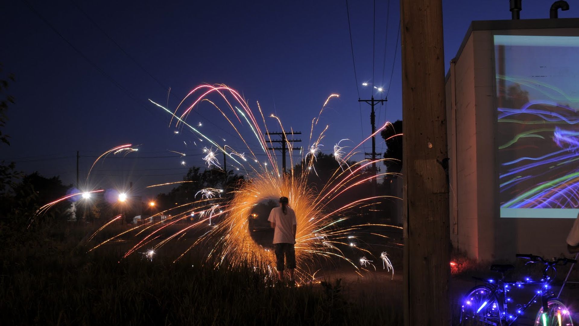 Person standing with back facing camera surrounded by a circular burst of bright orange and red fireworks against a deep blue night sky with light trails and a bicycle decorated with purple lights nearby.