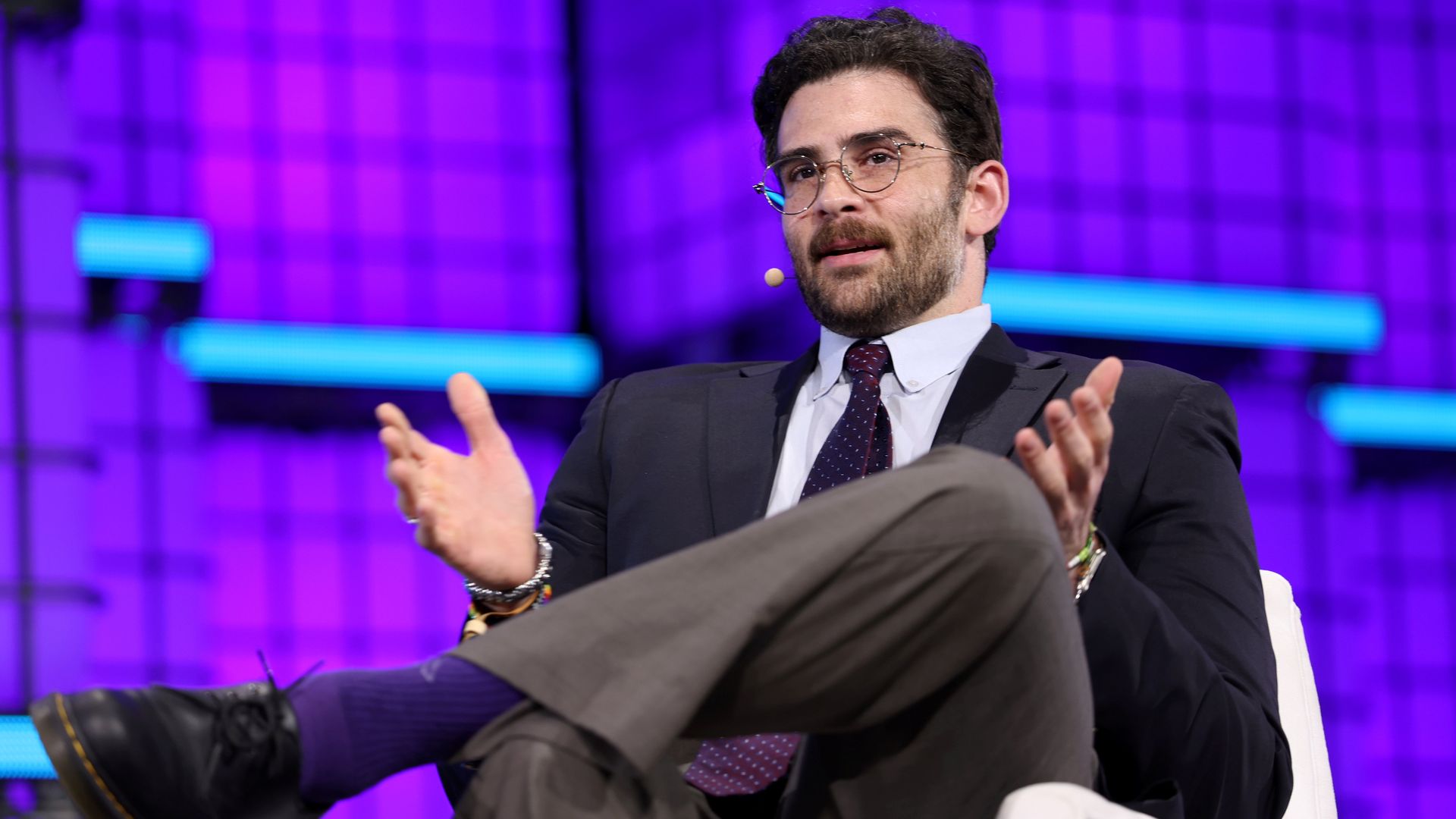Bearded man in a dark suit and maroon tie sits on a white chair on a purple-lit stage, wearing glasses and a headset microphone, gesturing with his hands.