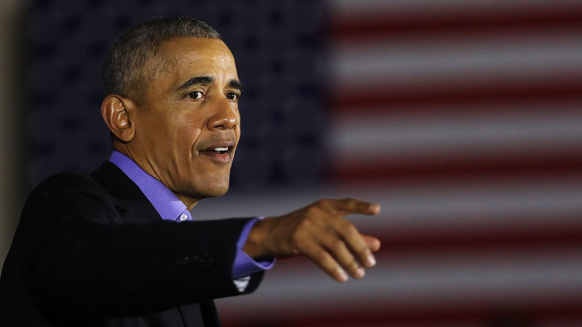 Barack Obama points, standing before a blurry American flag.
