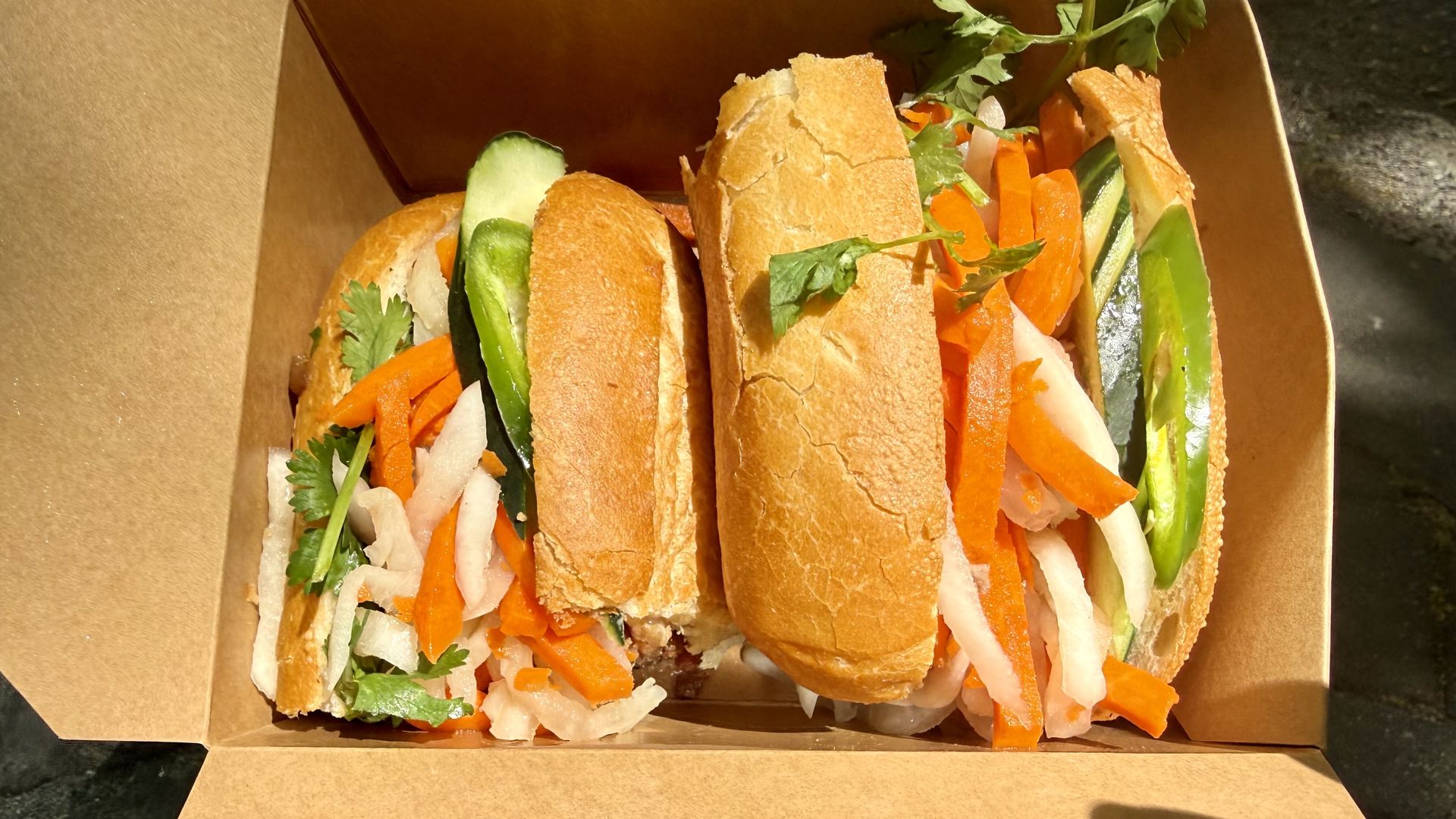 Two crusty baguette sandwiches in a brown takeout box filled with shredded carrot, cucumber strips, onion slices, and cilantro, with fresh greens visible in warm light.