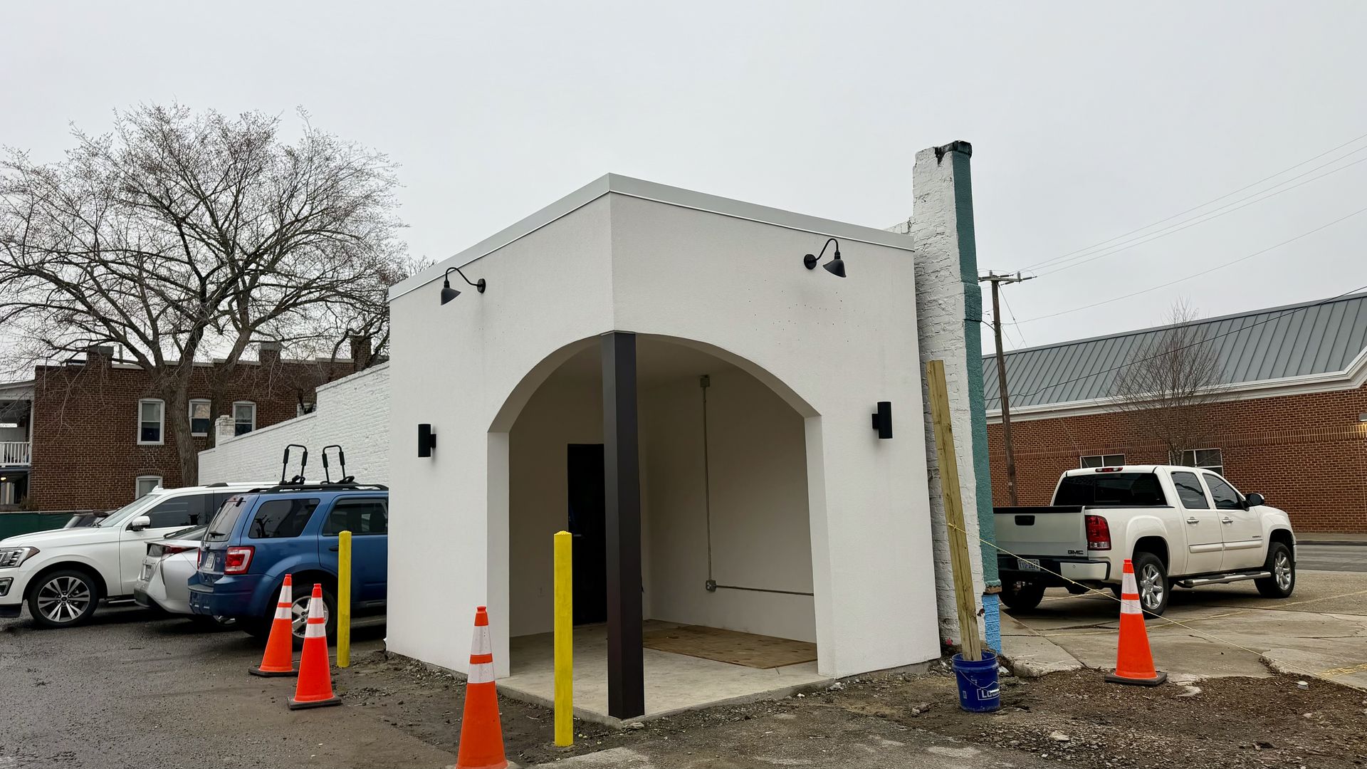 Small white building with an arched open entrance, brown support beam, black lights, and orange traffic cones around it, parked cars nearby, gray overcast sky.