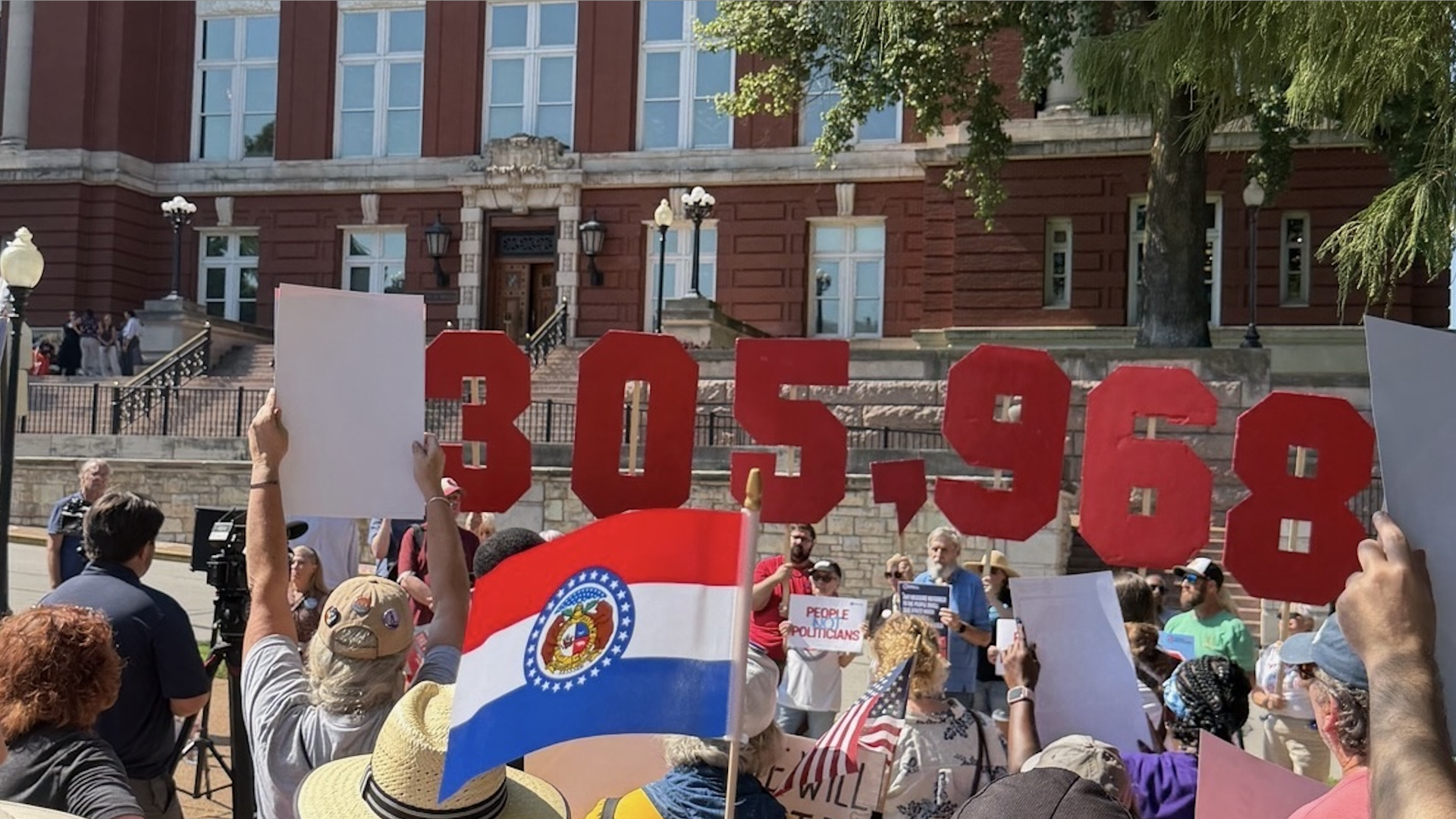 Crowd of protesters outdoors in front of a red-brick government building, holding large red letters spelling SCOTUS, with signs, flags, and people wearing hats in the foreground.