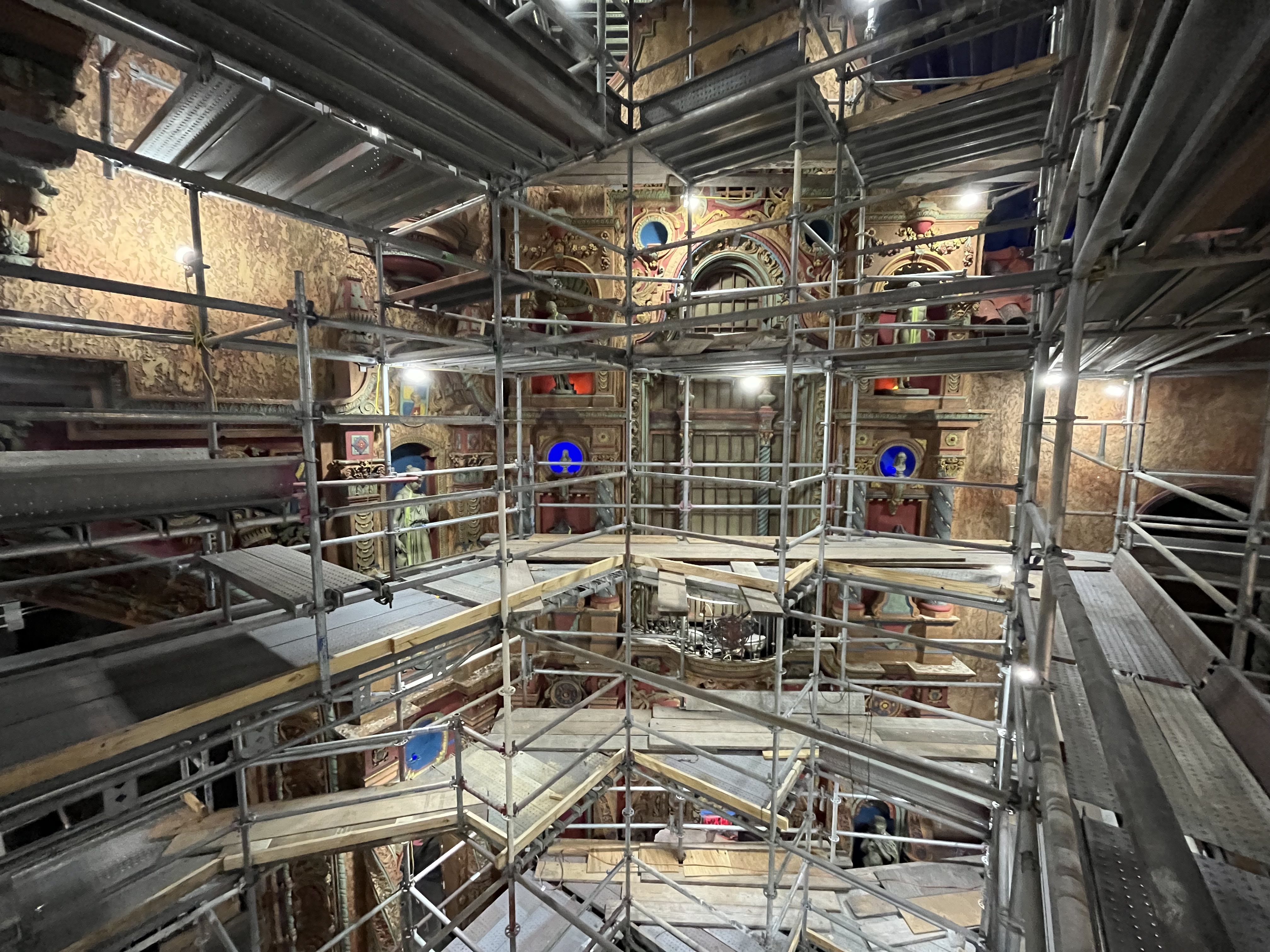 Interior of a historic space under dense metal scaffolding; ornate gold arches and murals are visible behind the lattice, warm lights, blue circular medallions, and wooden planks on the floor.