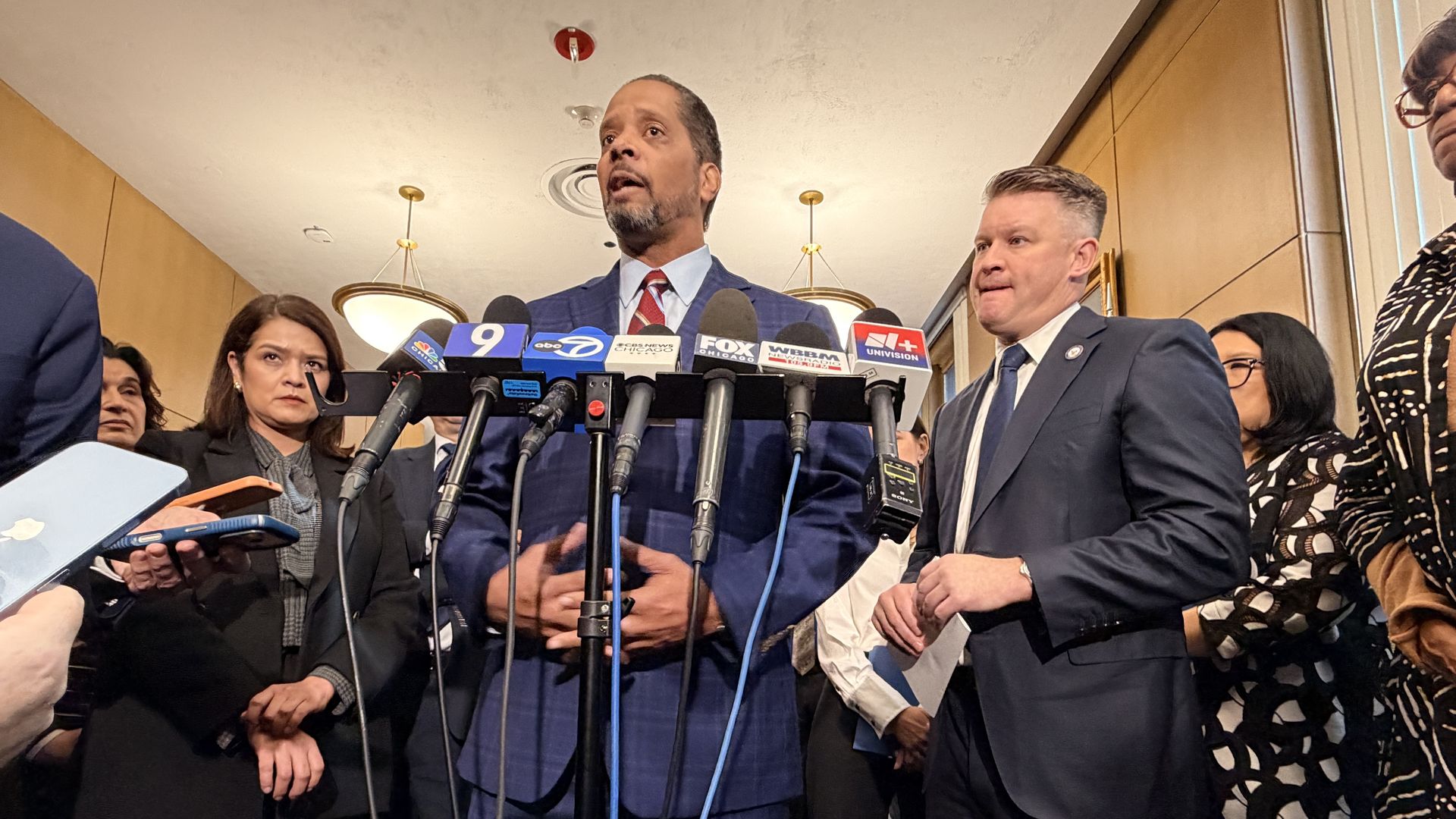 Man in a blue suit and red tie speaks at a podium cluttered with microphones from multiple news outlets; reporters surround him in a hallway during a press conference.