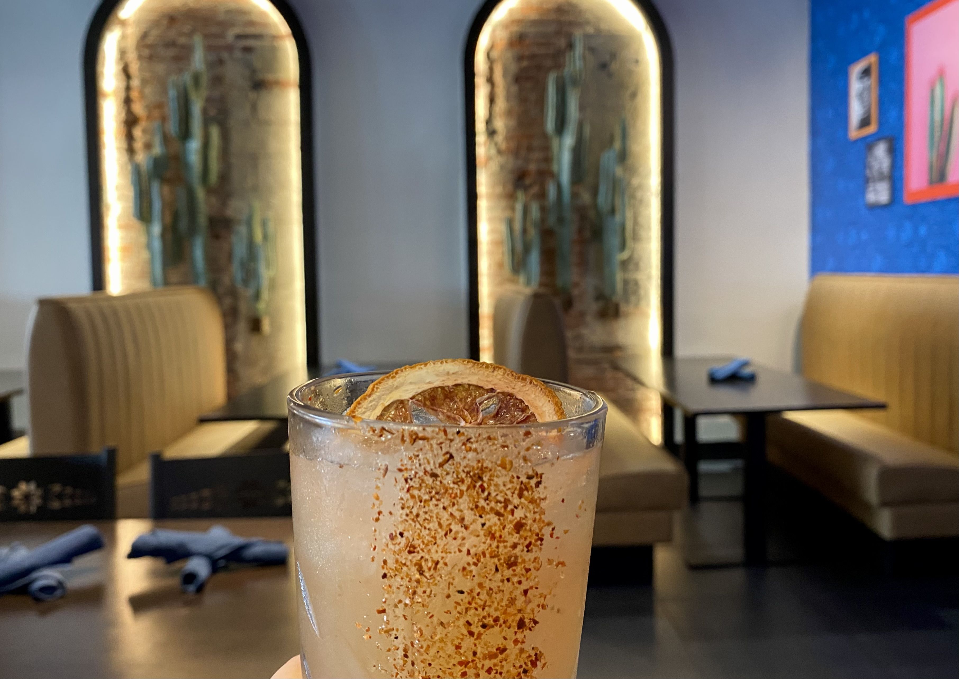 Light pink cocktail in a clear glass garnished with a dried citrus slice and chili powder, held in a modern restaurant with tan booth seating, blue walls, and cactus-themed decor.