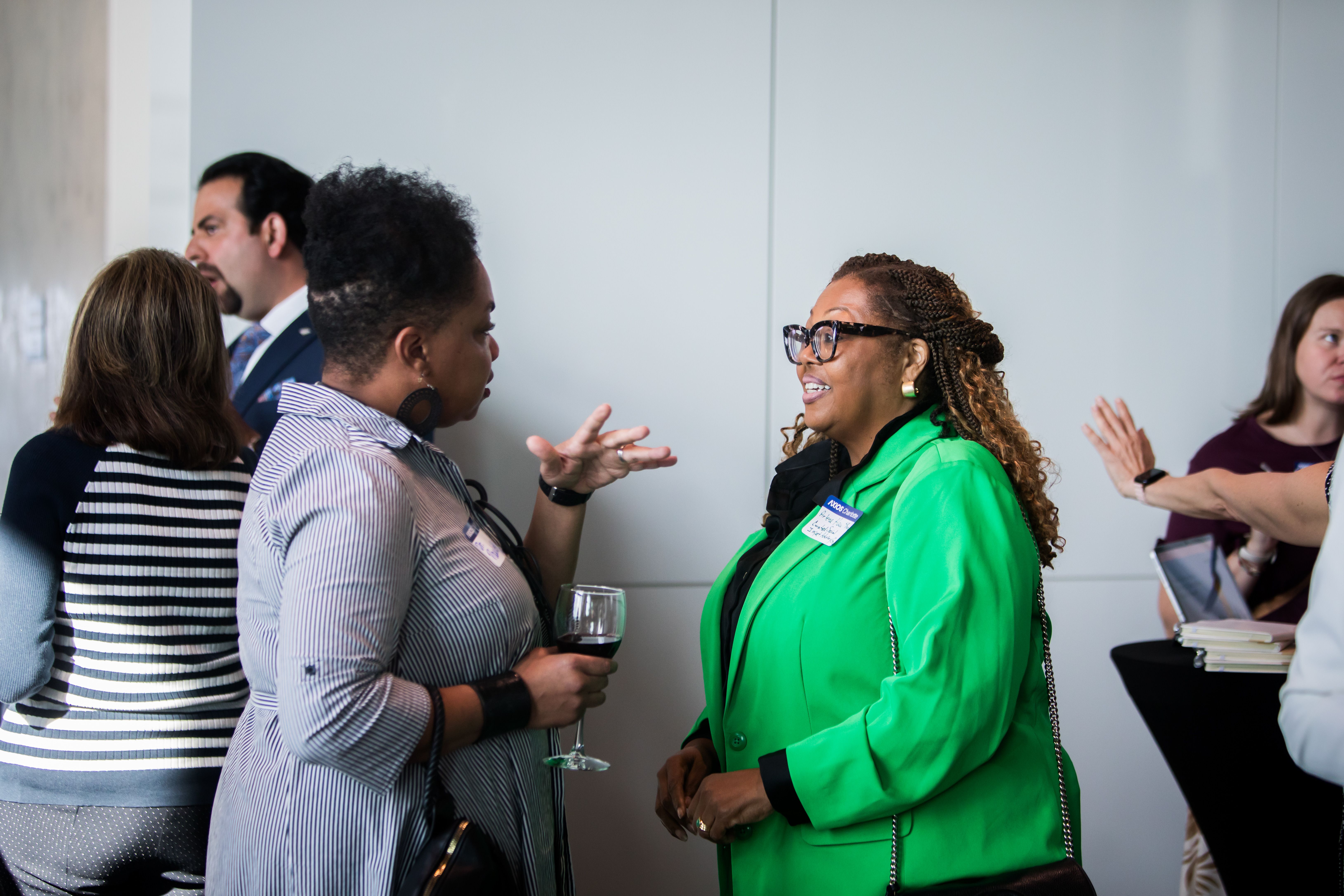 People mingle at a social event by a light gray wall: a woman in a bright green blazer chats with a striped-shirt woman, holding a wine glass; others stand nearby with name badges.