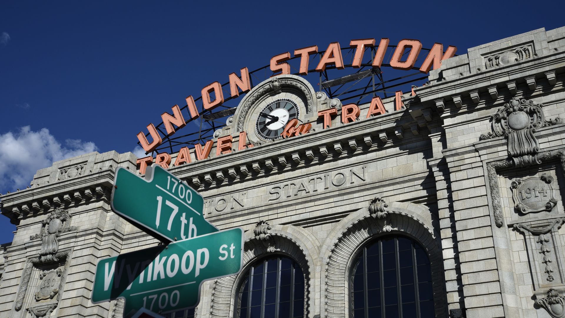 A large, classic building with the an orange sign reading UNION STATION, with two street signs in front of it. 