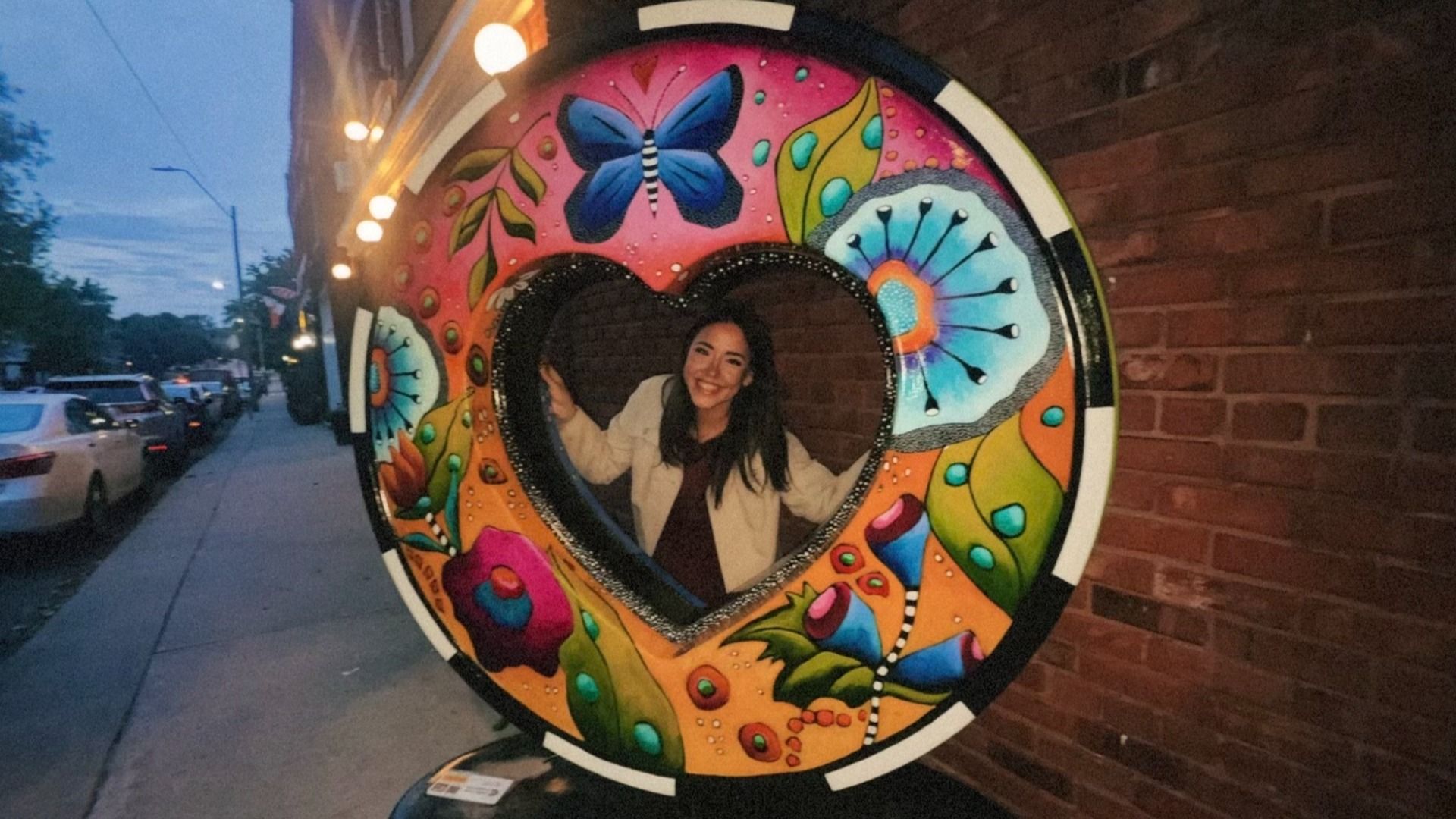 A smiling woman poses inside a colorful circular mural with a heart-shaped opening, on a brick wall by a sidewalk; pink background with blue butterfly and floral patterns at dusk.