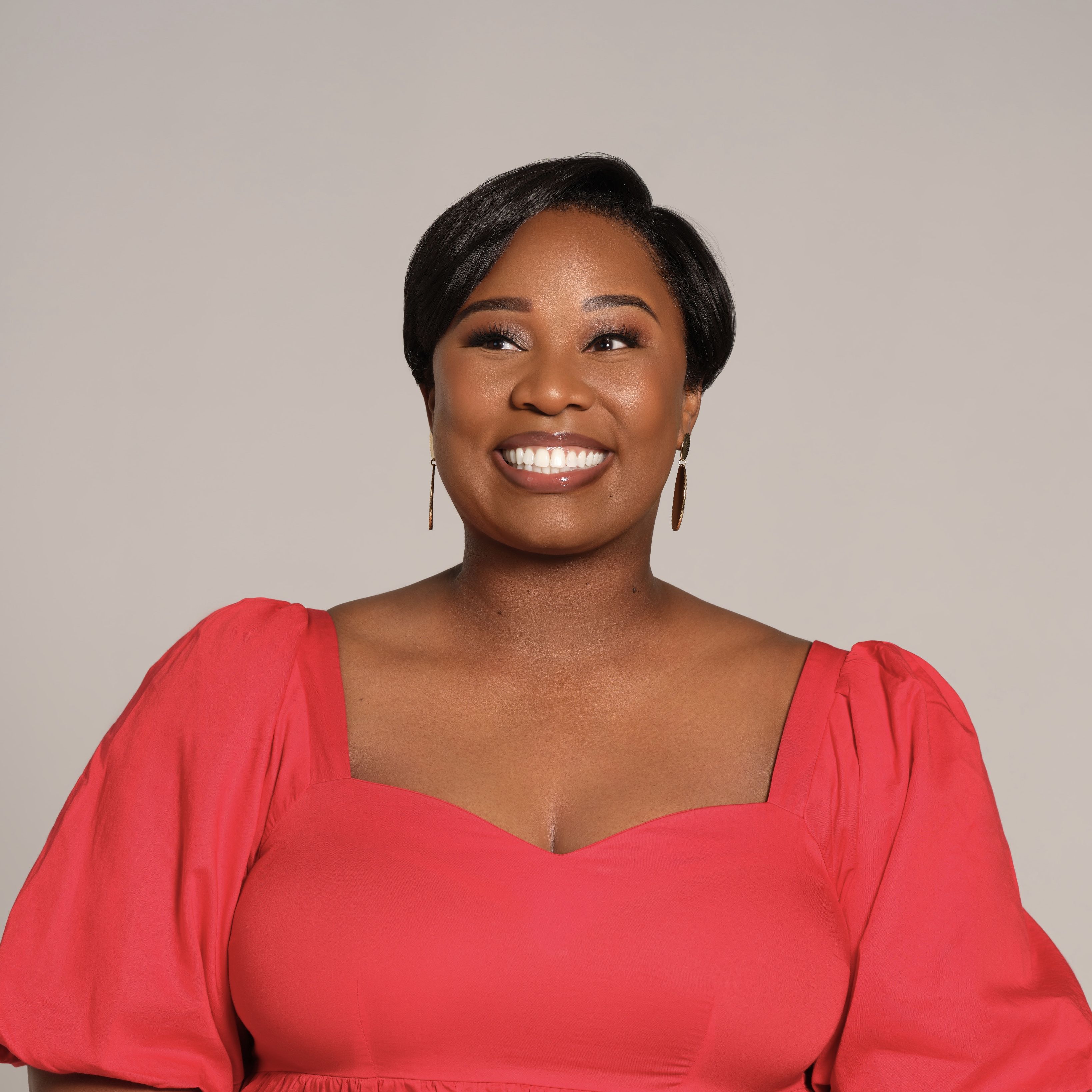 Close-up of a smiling Black woman in a coral pink dress with puff sleeves. Short black hair, bold makeup, gold earrings, neutral beige background.