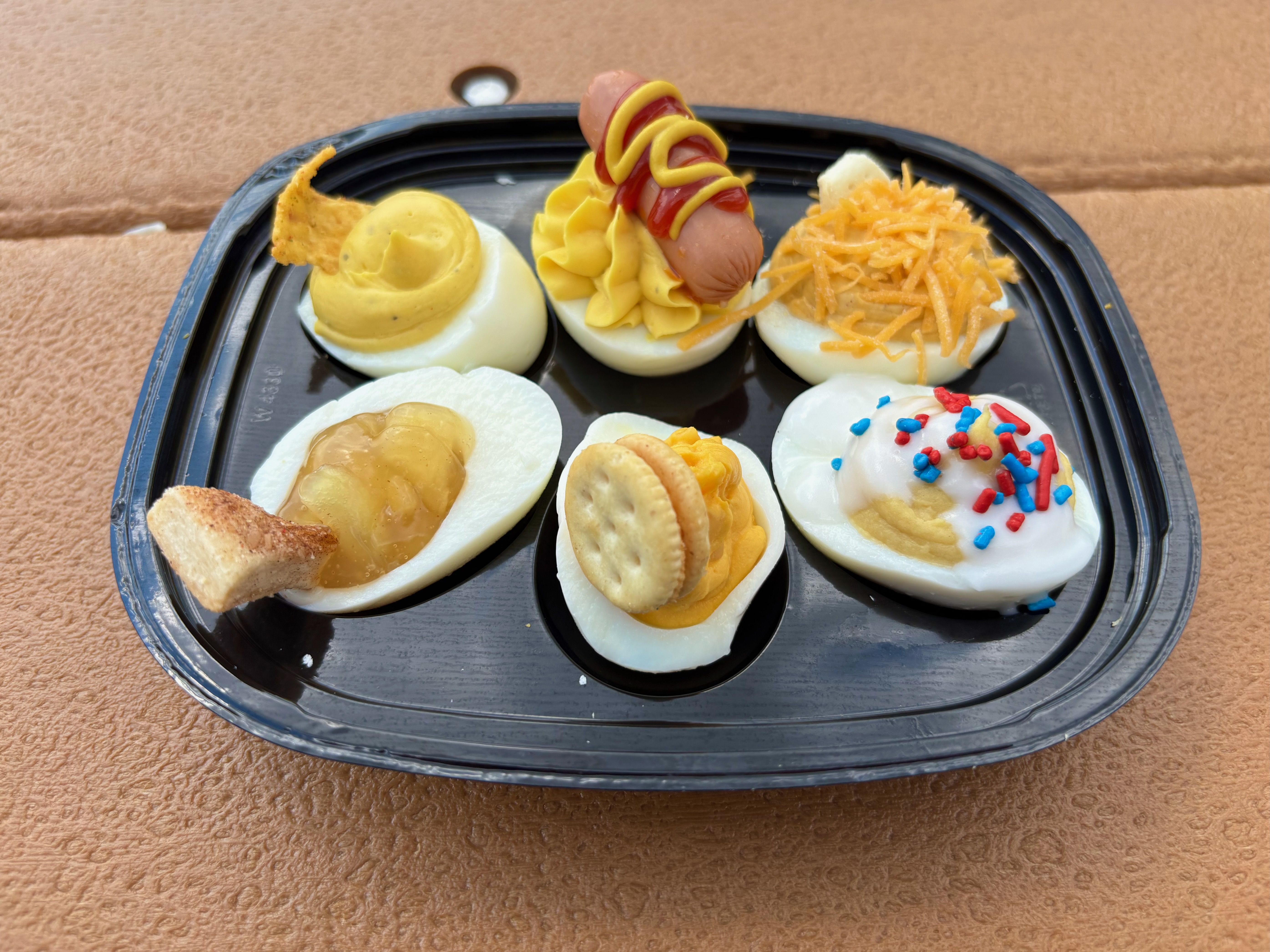 Tray of deviled eggs on a black plate with assorted toppings: a sausage-wrapped egg with mustard, cheese-topped eggs, and a sprinkles-covered egg.