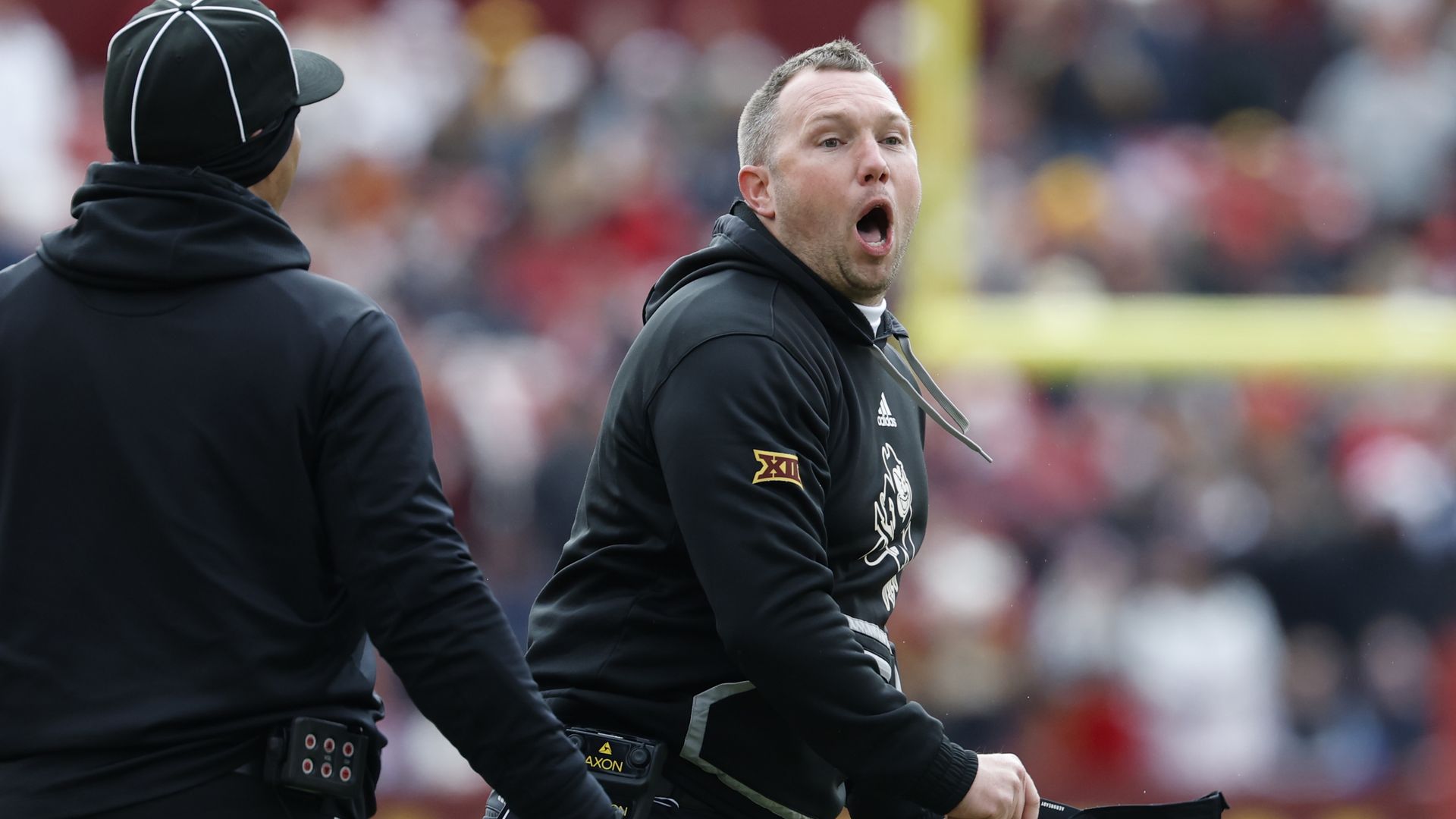 Two men in black sportswear on a football field, one facing away, the other holding a black cap and headset with mouth open, wearing a black hoodie with a Big 12 patch.