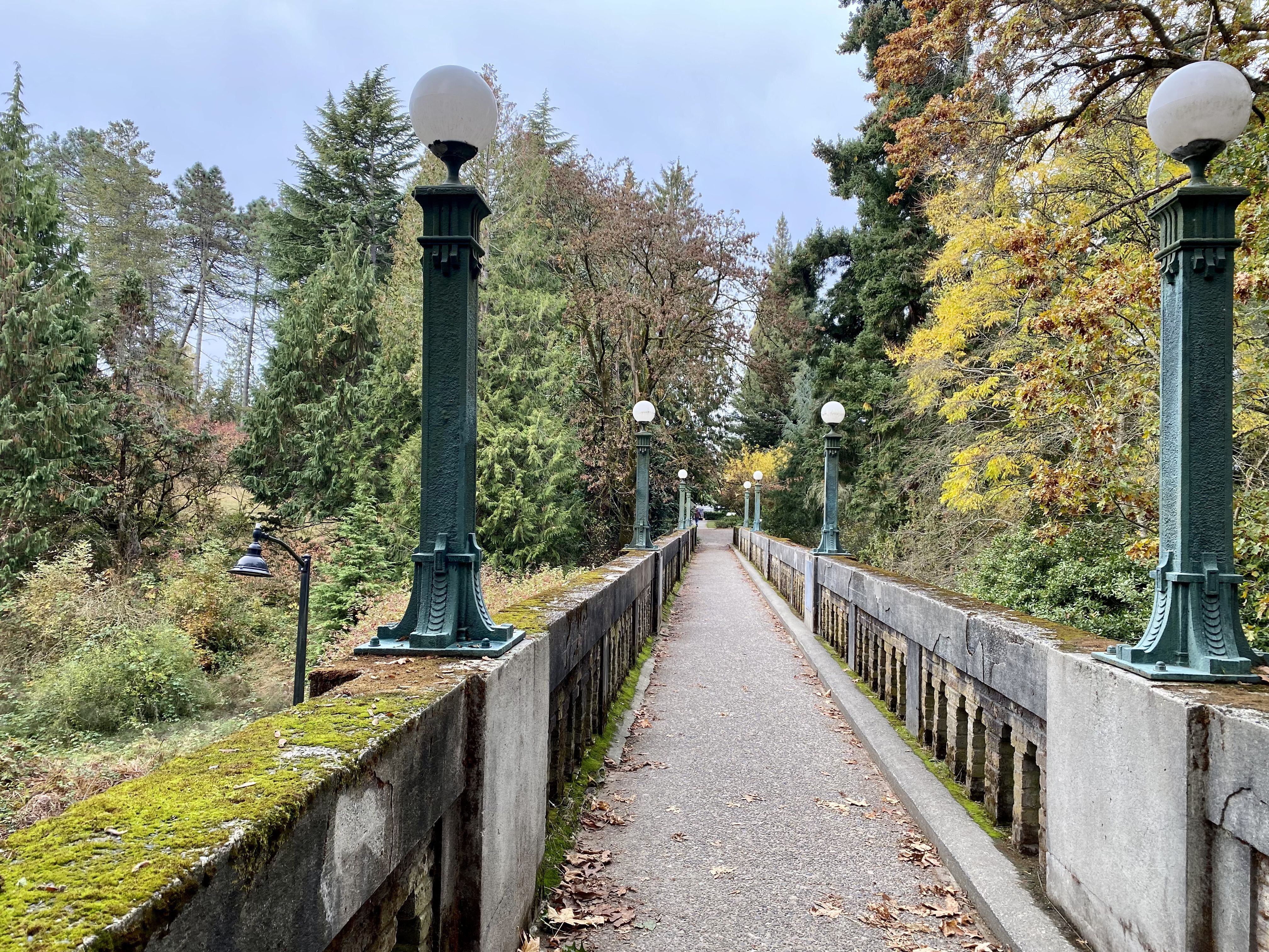 A bridge with some moss on it and patina covered lightposts with trees of different colors surrounding it. 