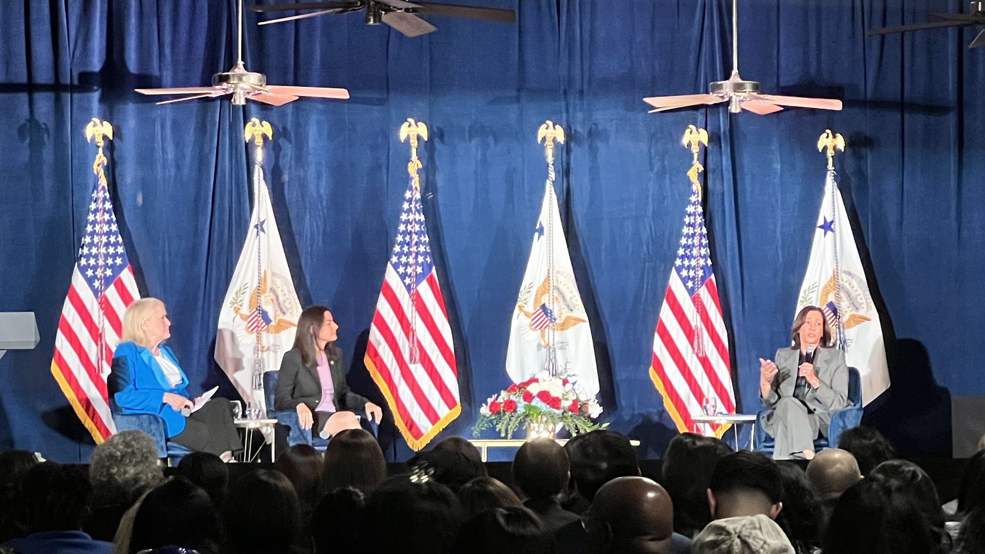 Photo of three women sitting on a stage, in front of multiple USA flags