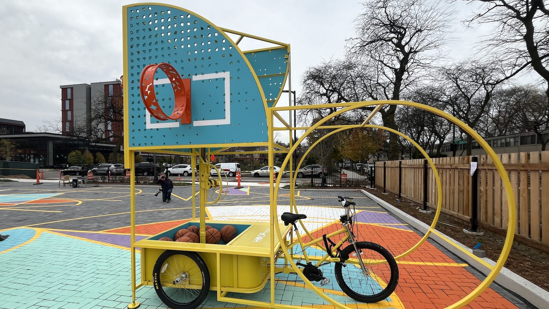 A large yellow tricycle that has a blue basketball backboard with an orange hoop and a basket with basketballs in it.