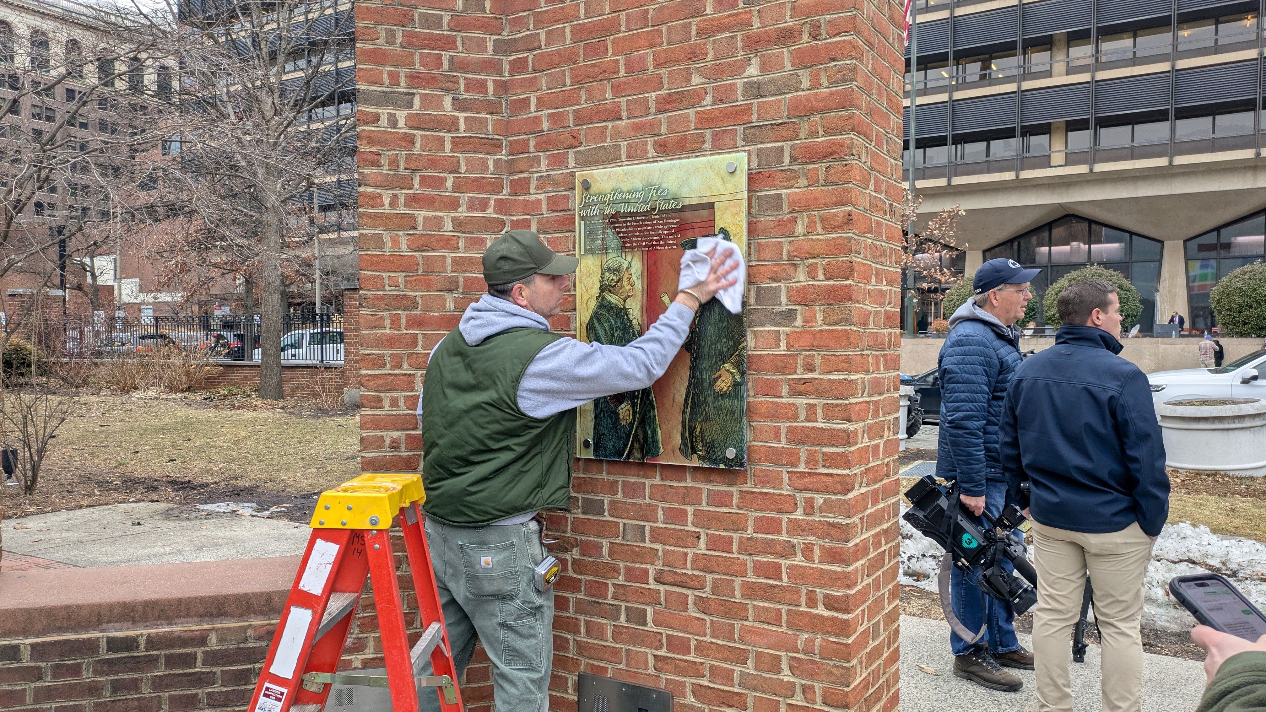 Man in green vest and cap cleaning a commemorative plaque on a red brick pillar, with two men and a camera crew nearby in an urban outdoor setting.