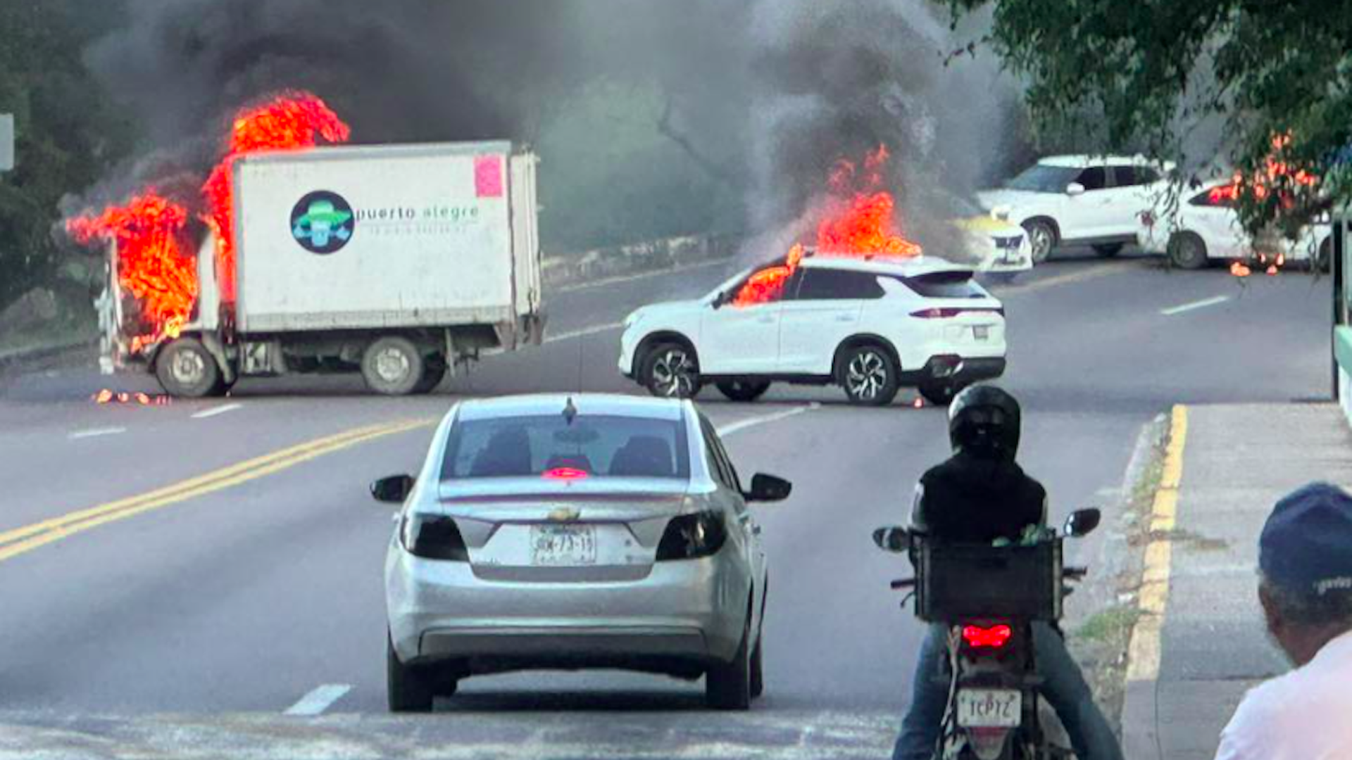 A white delivery truck and a white SUV are on fire, emitting black smoke, blocking a road. Several other vehicles and a person on a motorcycle are nearby.