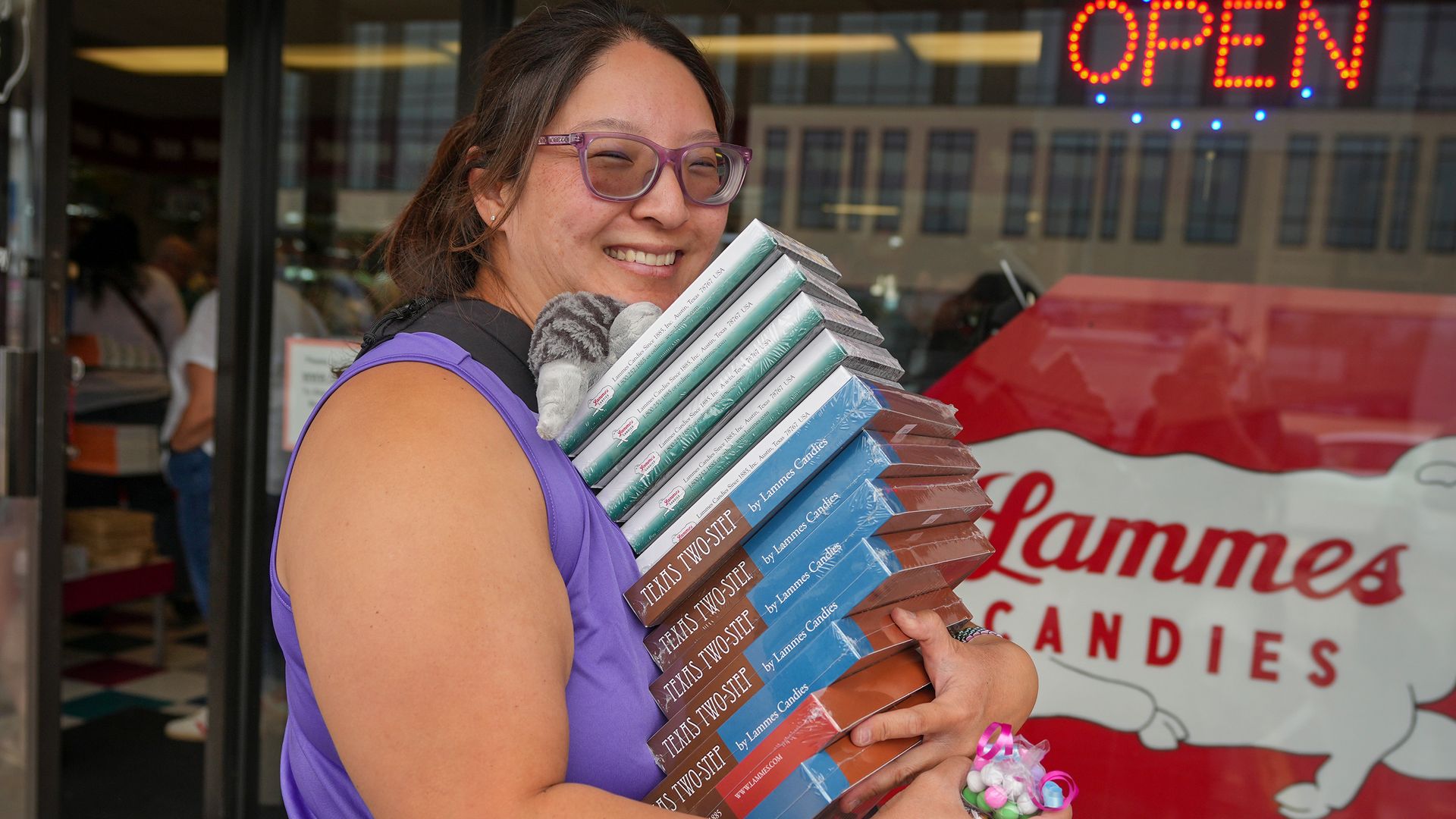 Smiling woman with purple glasses and a purple top holds a tall stack of boxed items labeled "Texas Two Step" outside a Lammes Candies storefront, with a bright OPEN sign in the background.