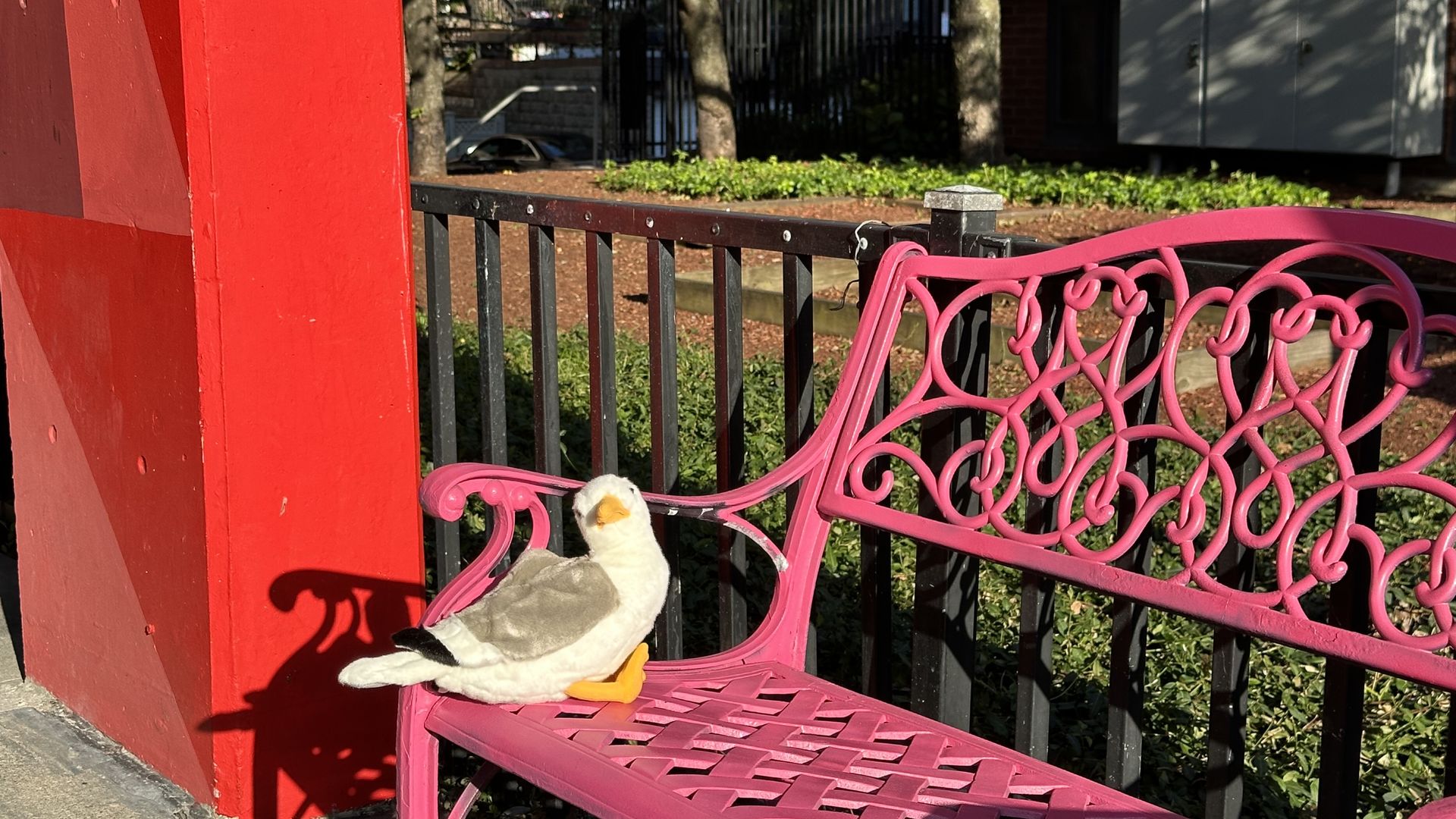 Townie, Axios Boston's plush gullscott, is sitting on a bink bench near a building beam that's painted red in an undisclosed location.
