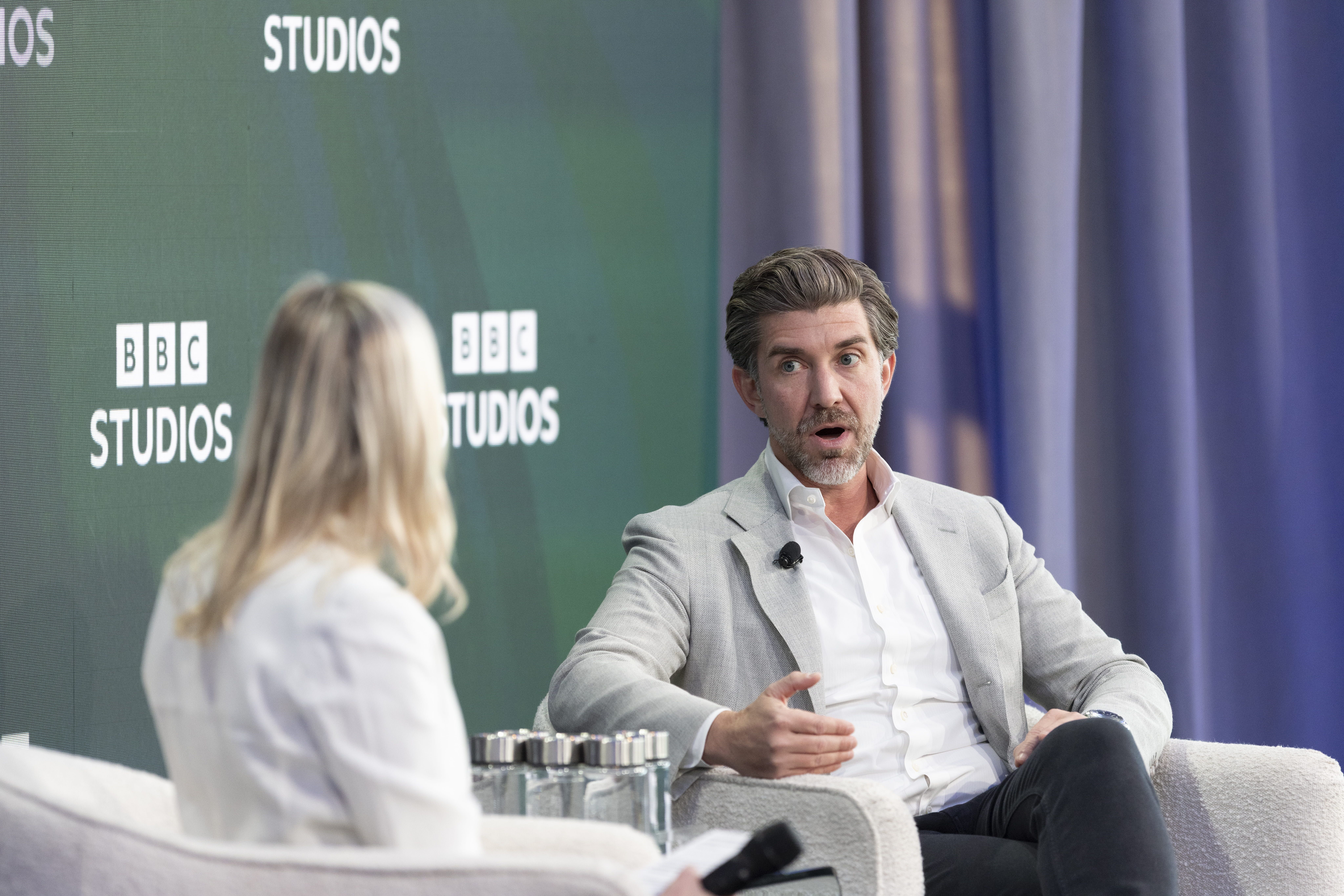 Two people in a TV studio: a man in a light gray suit speaks into a microphone while seated, facing a person with their back to the camera; BBC Studios logos on a green backdrop with blue curtains.